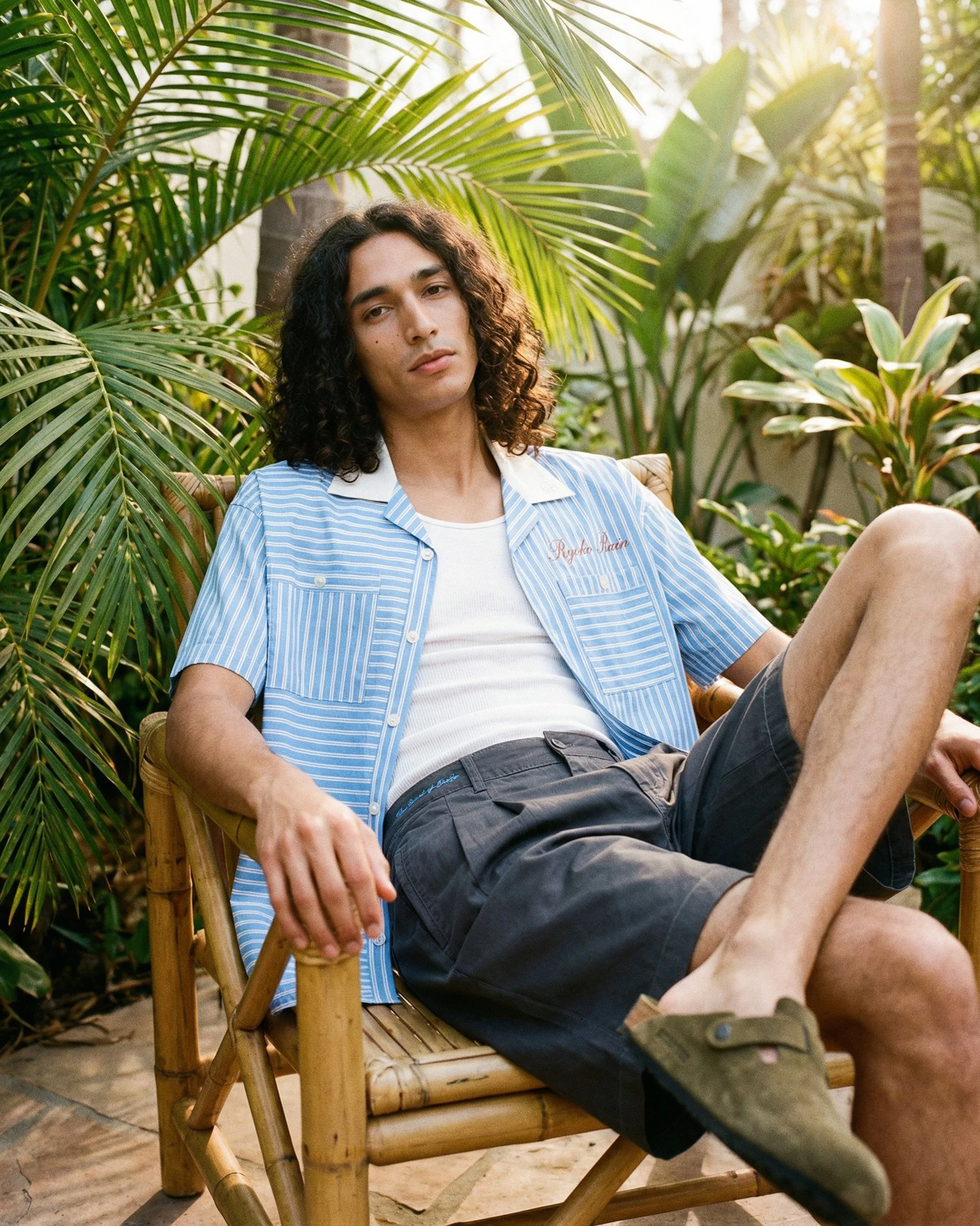 A young man with long curly dark hair sitting in a bamboo chair outdoors surrounded by lush green tropical plants. He is wearing a blue and white striped shirt over a white T-shirt and dark shorts, relaxing with a calm expression.