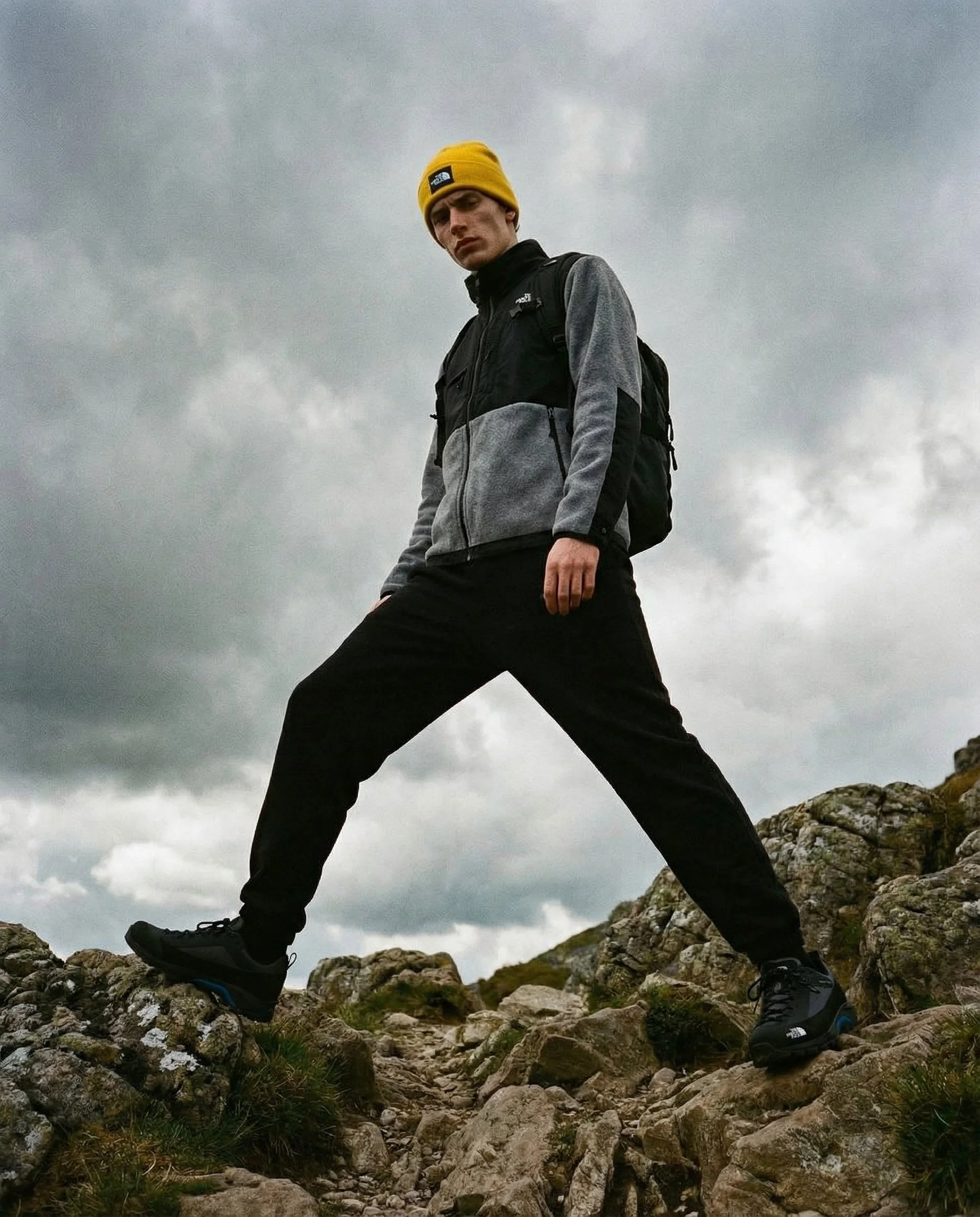 A man wearing a yellow beanie, black North Face jacket, and black hiking boots stands with legs apart on rocky terrain during daytime with cloudy sky.