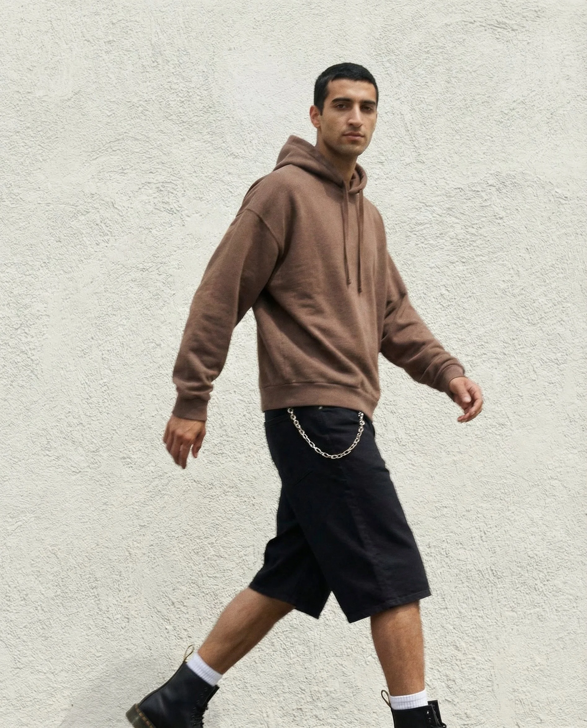 A young man with short dark hair wearing a brown hoodie, black shorts with a chain, white socks, and black boots, walking against a textured white wall.