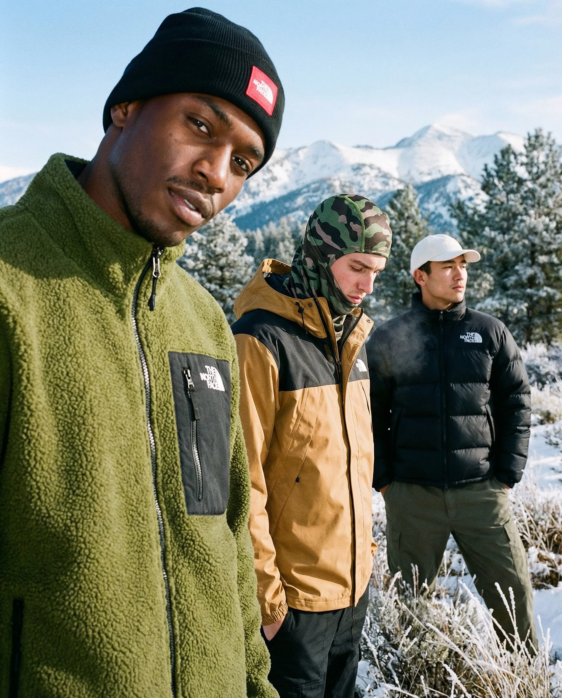 Three young men standing outdoors in a snowy mountain landscape. The man in the foreground wears a black beanie and a green fleece jacket. The other two men wear a camouflage hooded jacket and a black puffer jacket with a white cap. All are dressed i