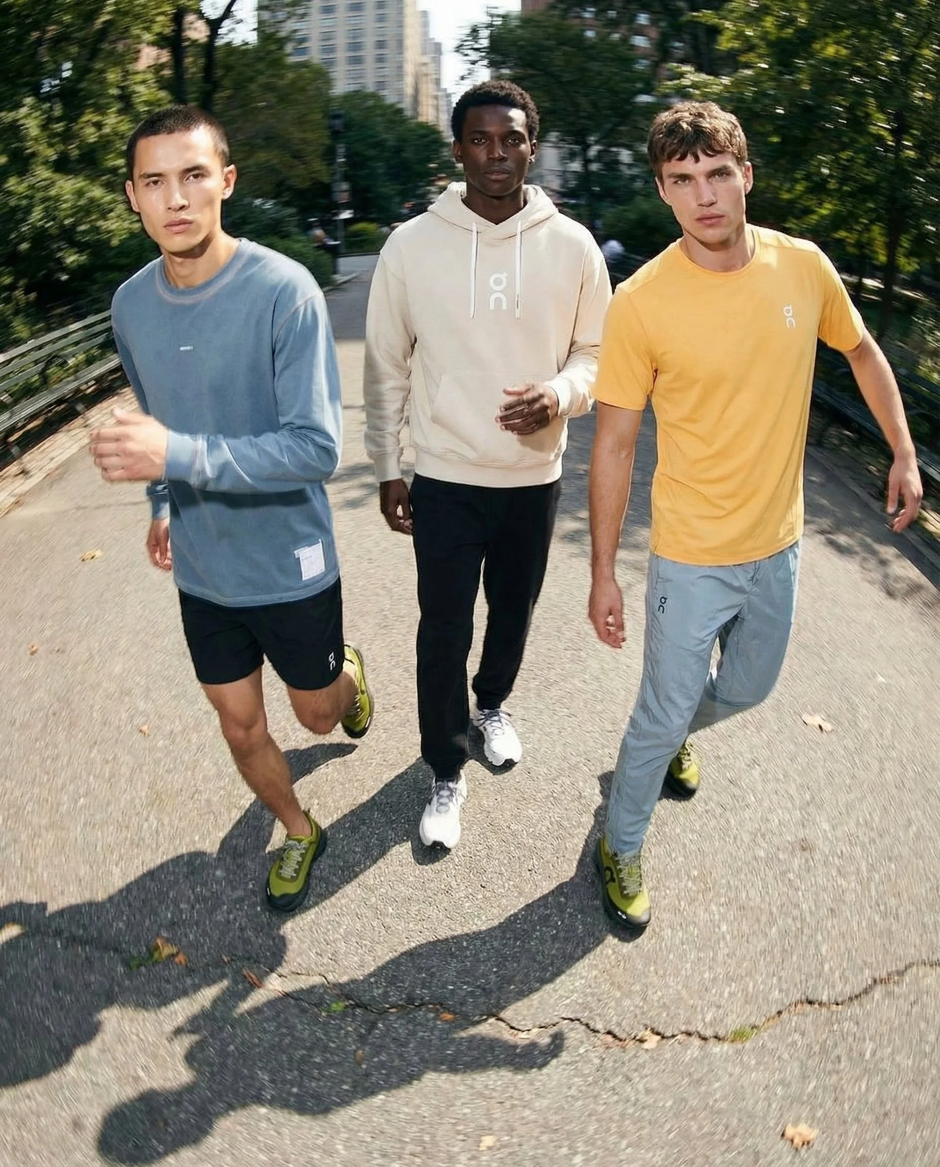 Three young men jogging on a sidewalk in a city park with trees and buildings in the background.