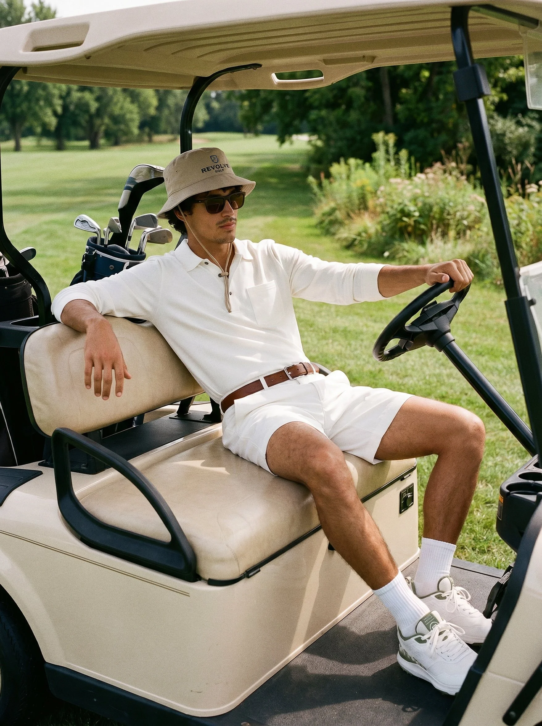 A man wearing sunglasses, a bucket hat, a white shirt, and shorts sitting relaxed in a golf cart on a golf course with trees and greenery in the background.