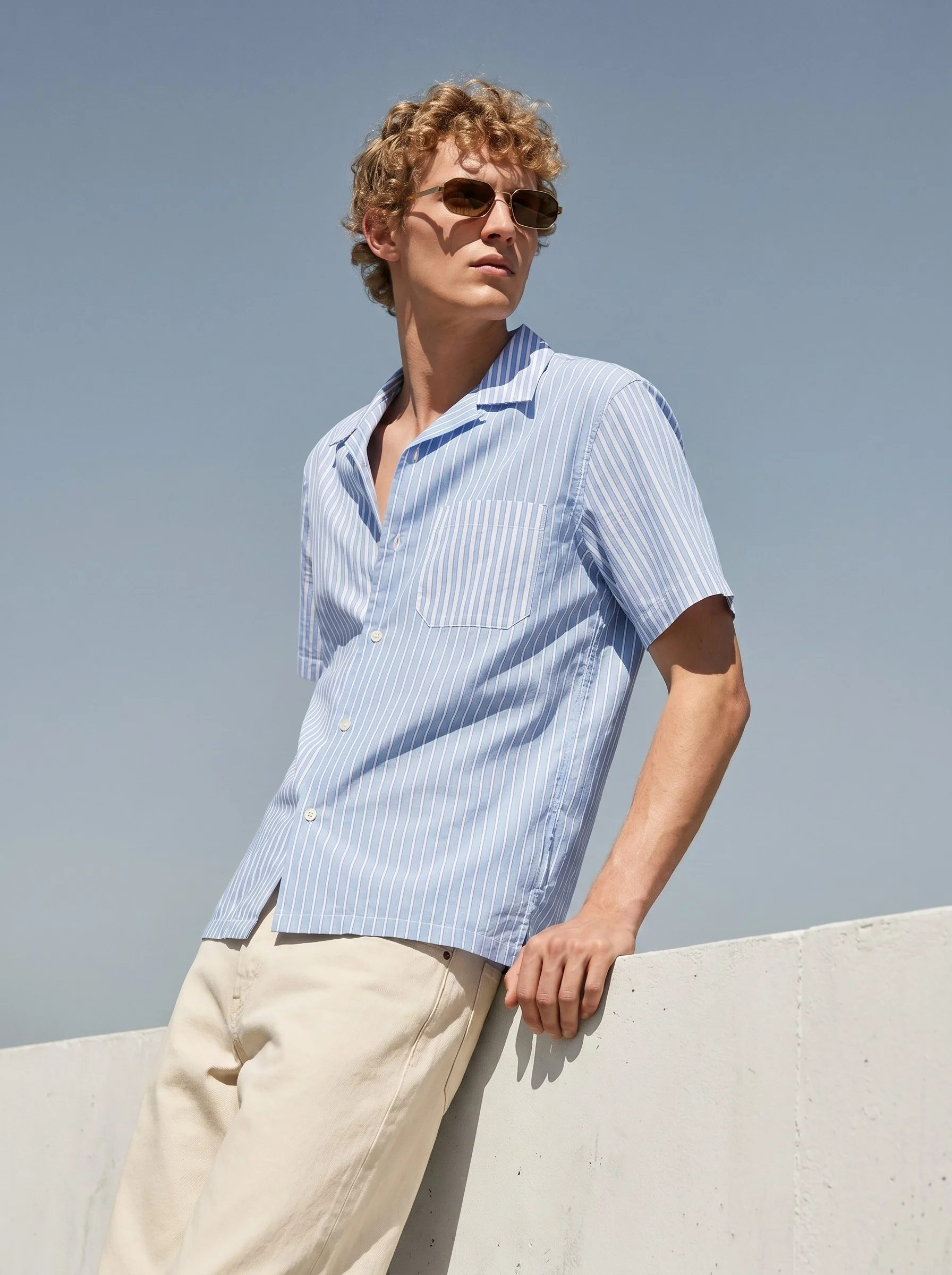 A young man with curly hair wearing sunglasses, a light blue striped short-sleeve shirt, and beige pants, standing outdoors against a pale wall, with a clear blue sky in the background.
