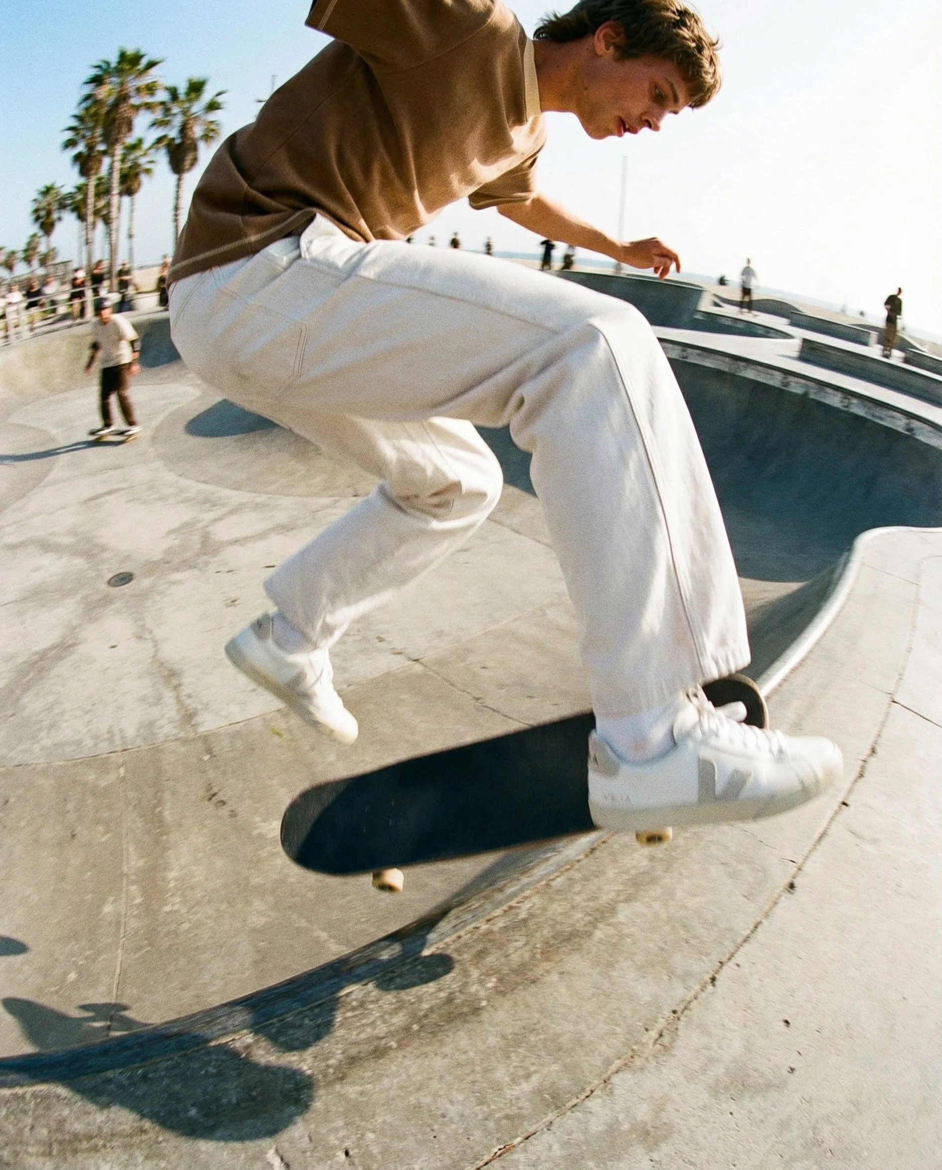 A young man skateboarding at the skatepark during late afternoon, wearing a brown t-shirt and white pants, with palm trees and other skateboarders in the background.