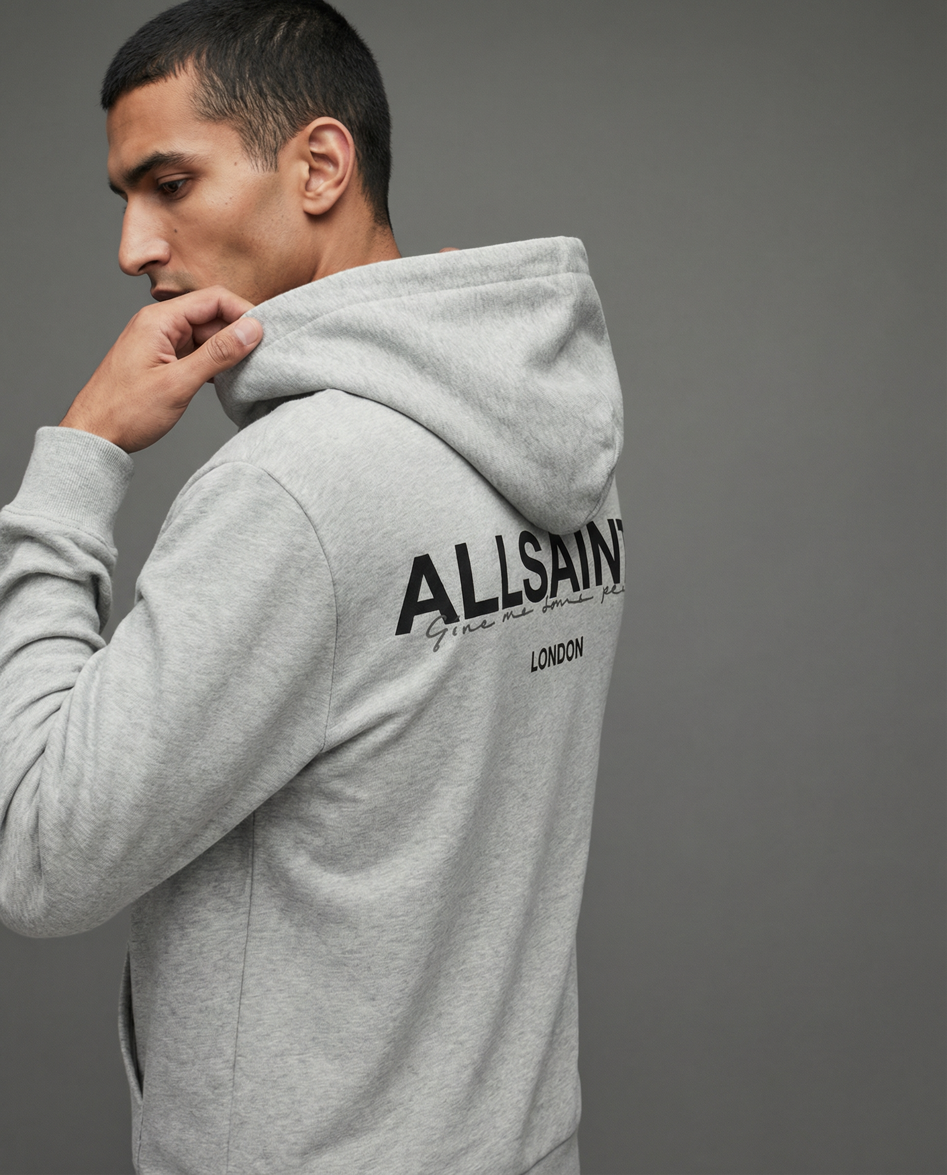 A man in a gray hoodie with text on the back standing against a gray background, looking down and touching the hood of his hoodie.