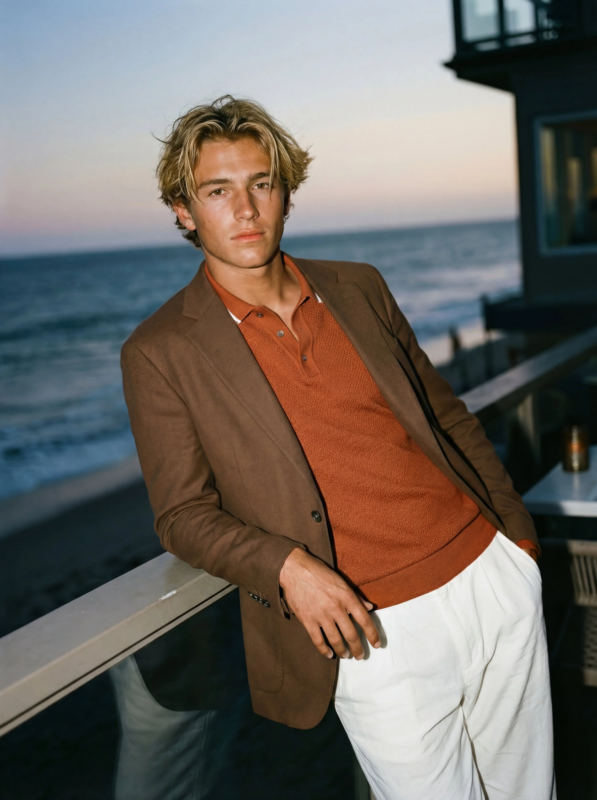 A young man with blond hair dressed in a brown blazer and orange polo shirt, standing on a balcony near the ocean at sunset.