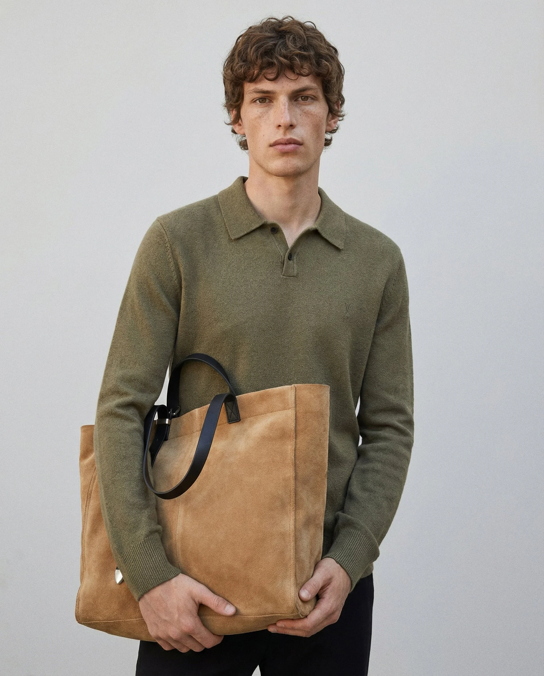 A person holding a tan paper shopping bag with black handles, wearing an olive green sweater, standing against a plain light gray background.
