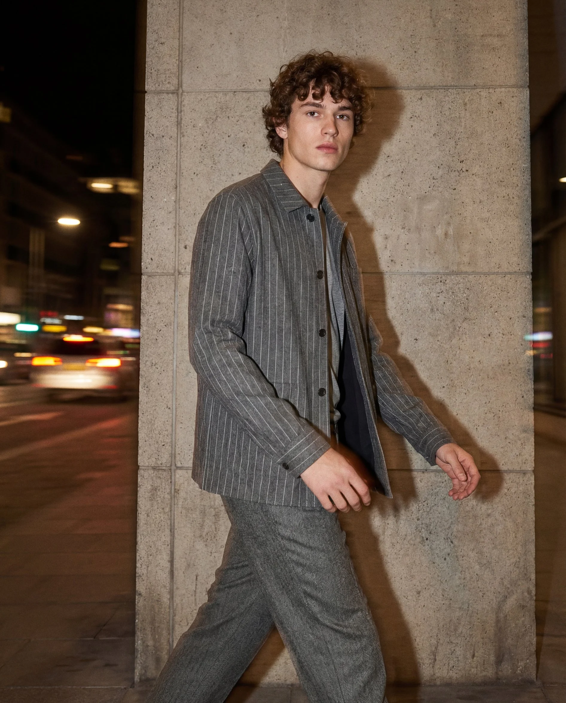A young man with curly brown hair and light skin leaning against a concrete wall at night, dressed in a gray pinstripe suit and gray pants, with blurred city lights in the background.