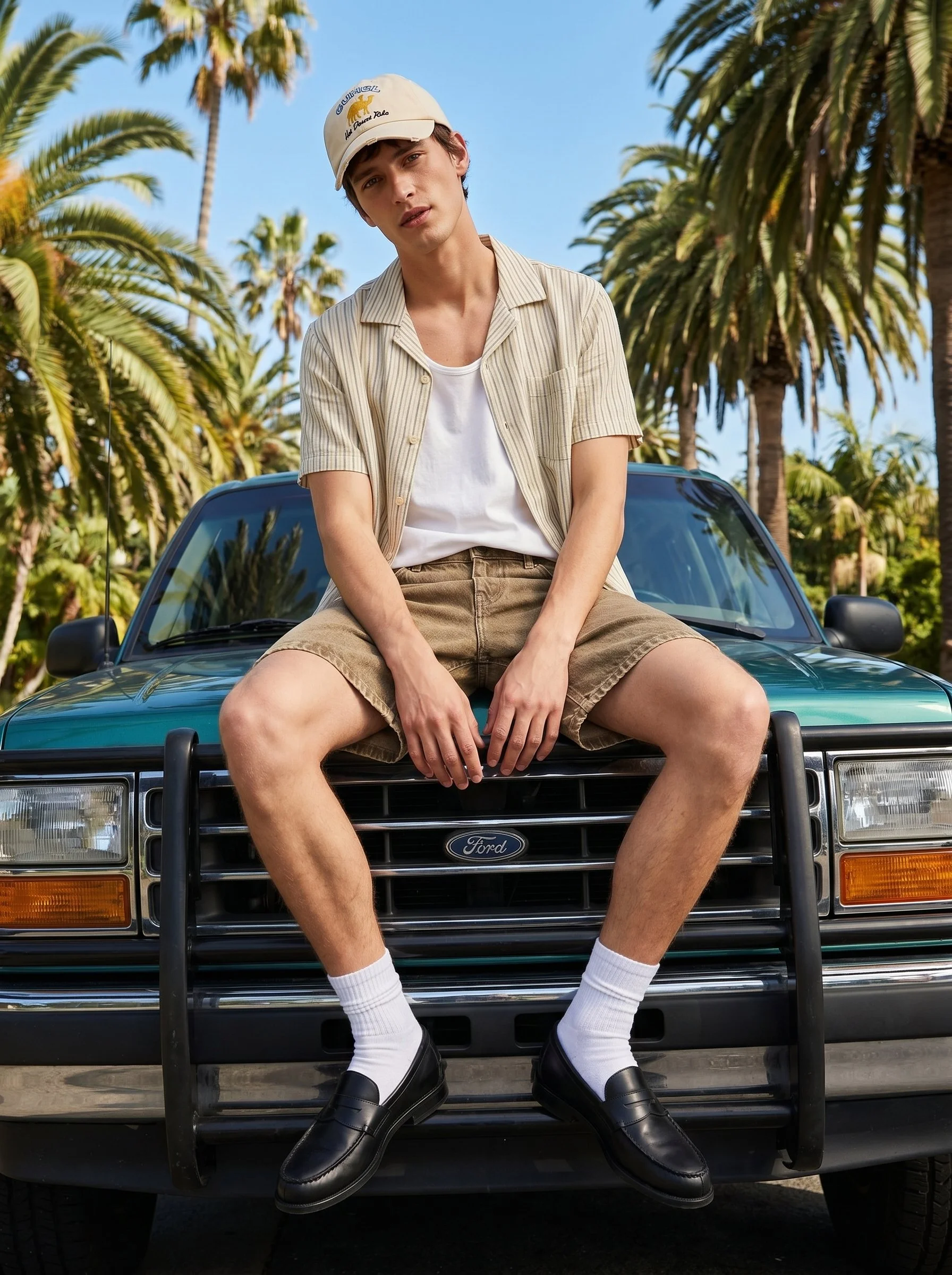 A young man wearing a cap, striped short-sleeve shirt, white t-shirt, beige shorts, white socks, and black loafers sitting on the hood of a green Ford vehicle with palm trees in the background.