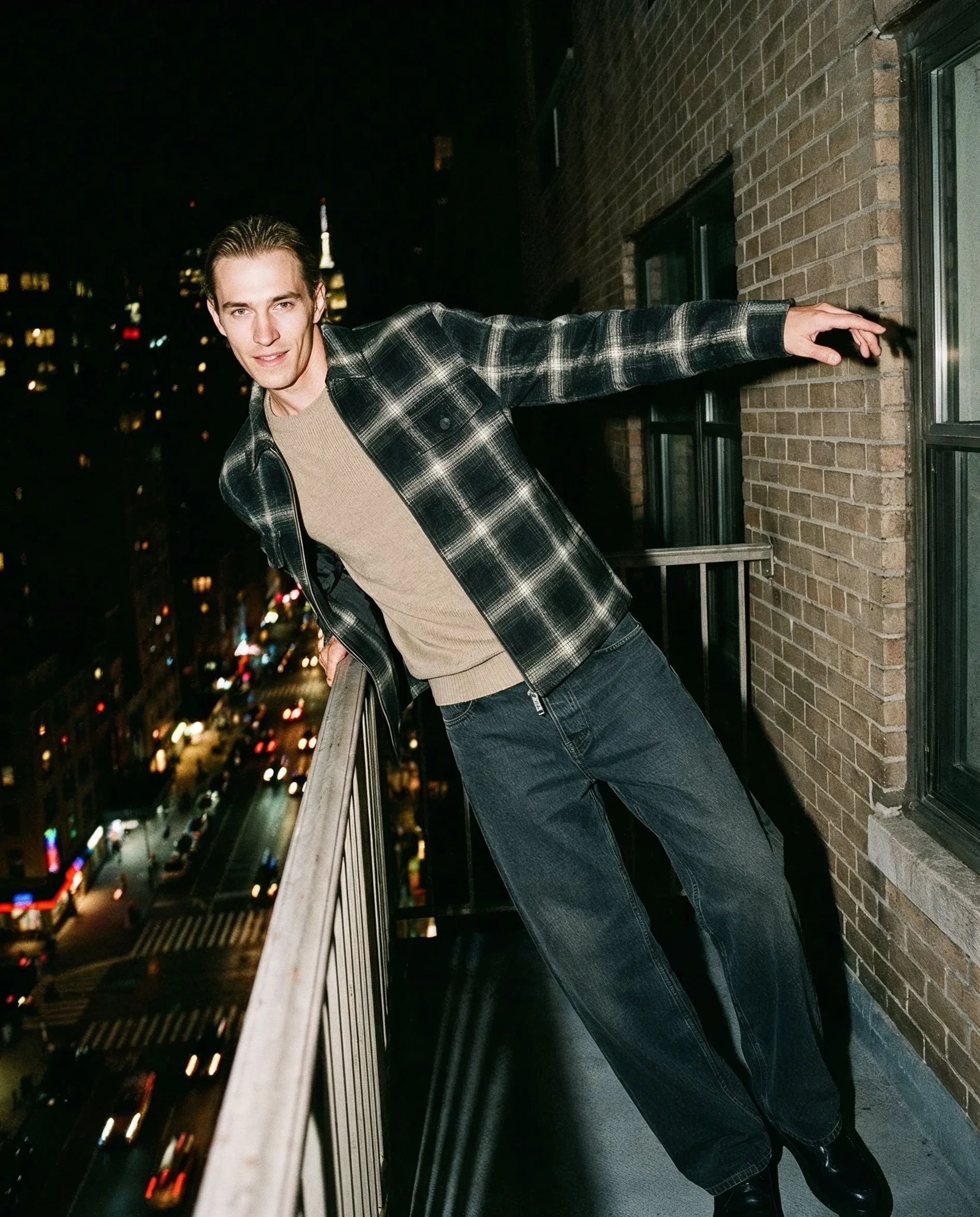 A young man with slicked-back hair wearing a black and white plaid jacket, beige sweater, and dark jeans, leaning on a brick building's fire escape railing at night, city lights blurred in the background.