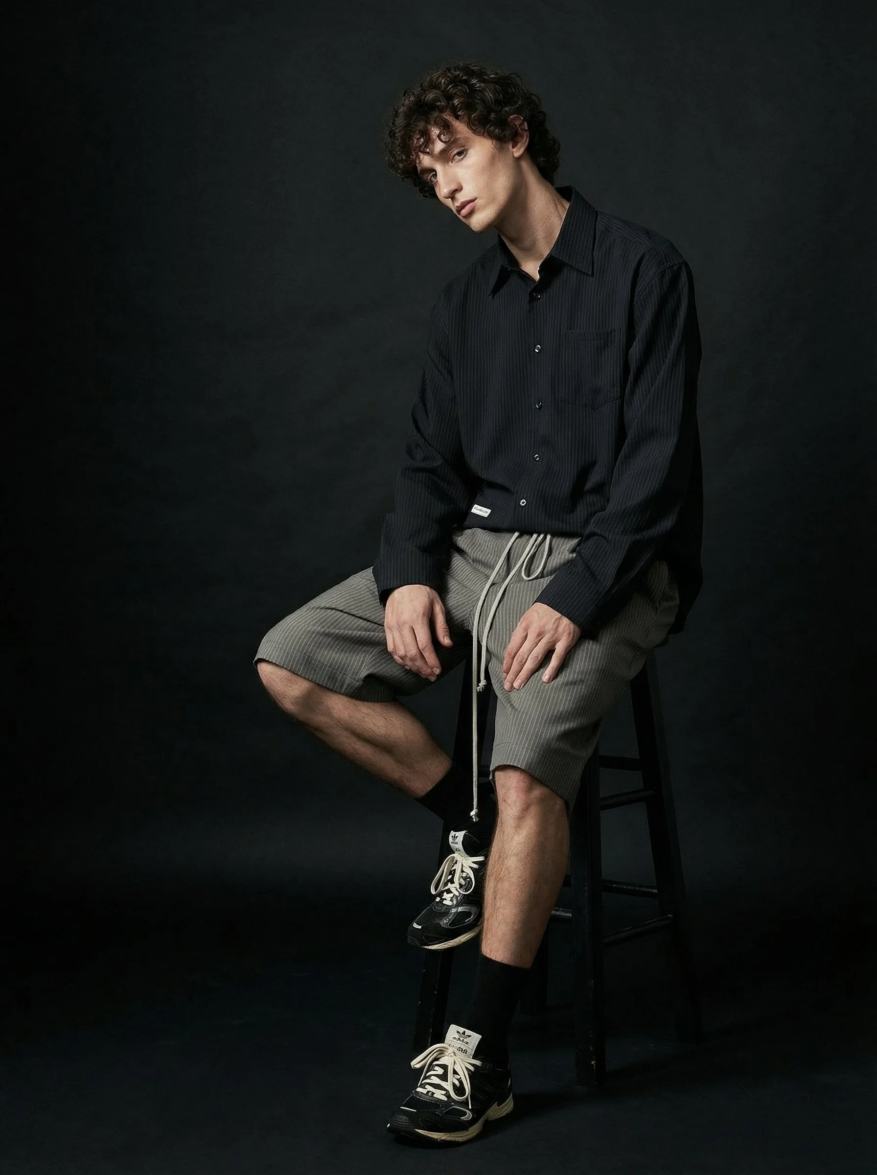 A young man with curly hair sitting on a black stool against a dark background, wearing a black shirt, gray shorts, black socks, and sneakers.