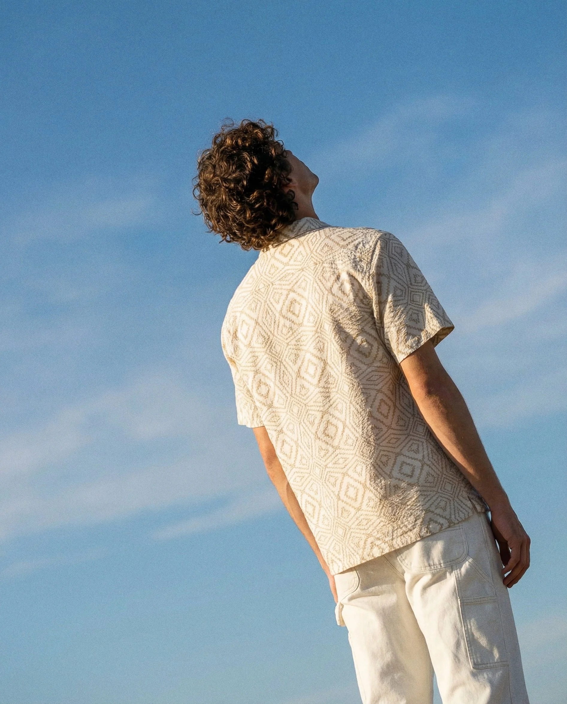 A person with curly hair wearing a beige patterned short-sleeve shirt and light-colored pants, standing outdoors against a blue sky.