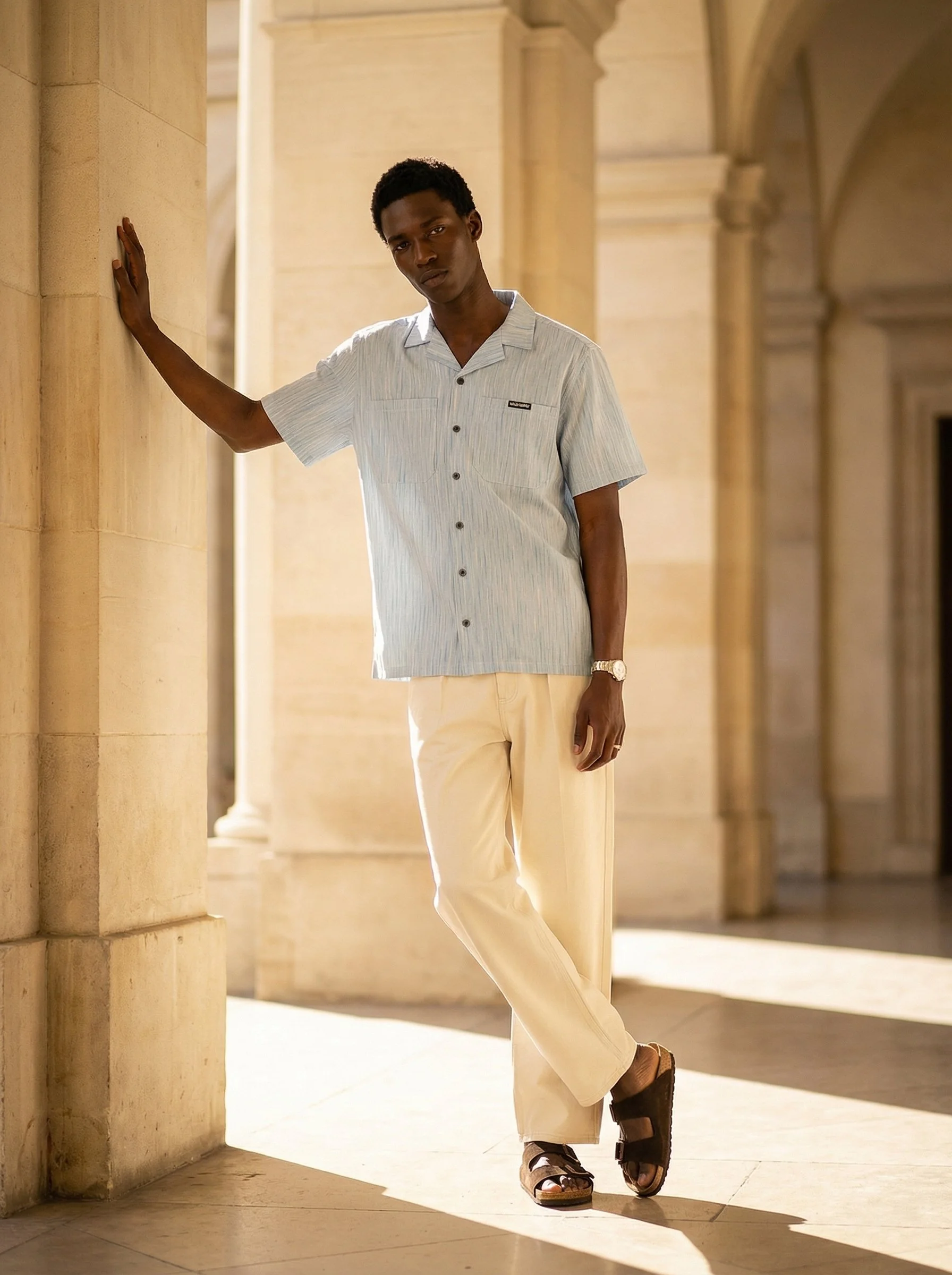 A young man with dark skin, short curly hair, wearing a light blue short-sleeved button-up shirt and beige wide-leg pants, stands indoors with his left hand on a stone wall and his right hand relaxed by his side, in a corridor with stone columns and 
