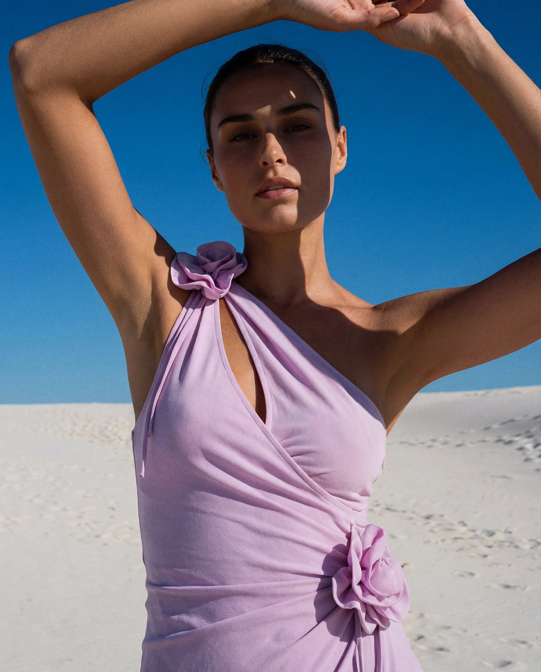 A woman with dark hair tied back, wearing a light purple wrap dress with a ruffle on one shoulder and a bow at the waist, standing on a white sandy beach under a clear blue sky, raising her arms above her head.