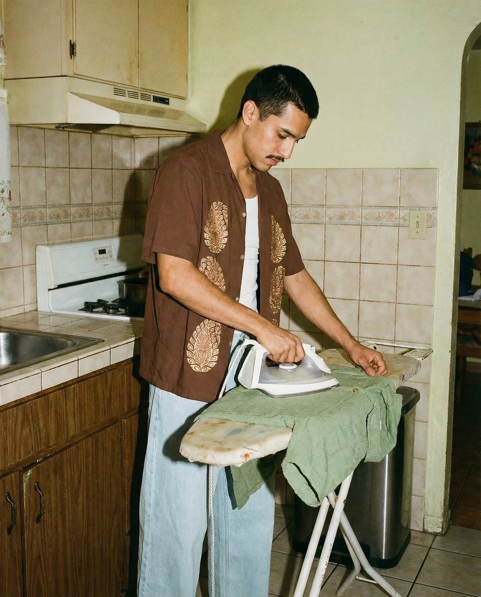 A man in a brown patterned shirt and light blue jeans is ironing clothes on an ironing board in a kitchen.