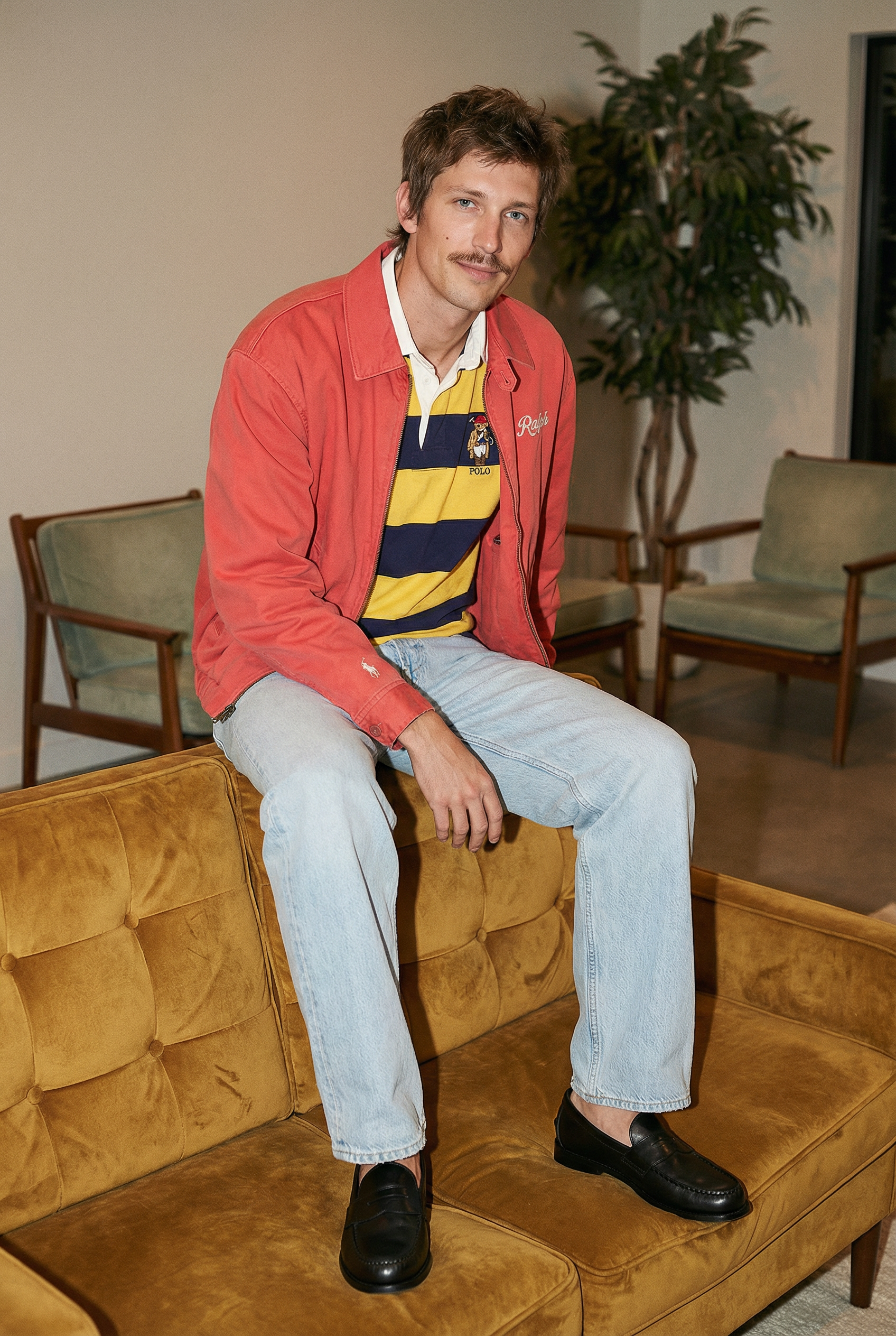 A man sitting on a vintage leather sofa in a living room, wearing a red jacket, a yellow and navy striped polo shirt, light blue jeans, and black loafers. There are chairs and a potted plant behind him.