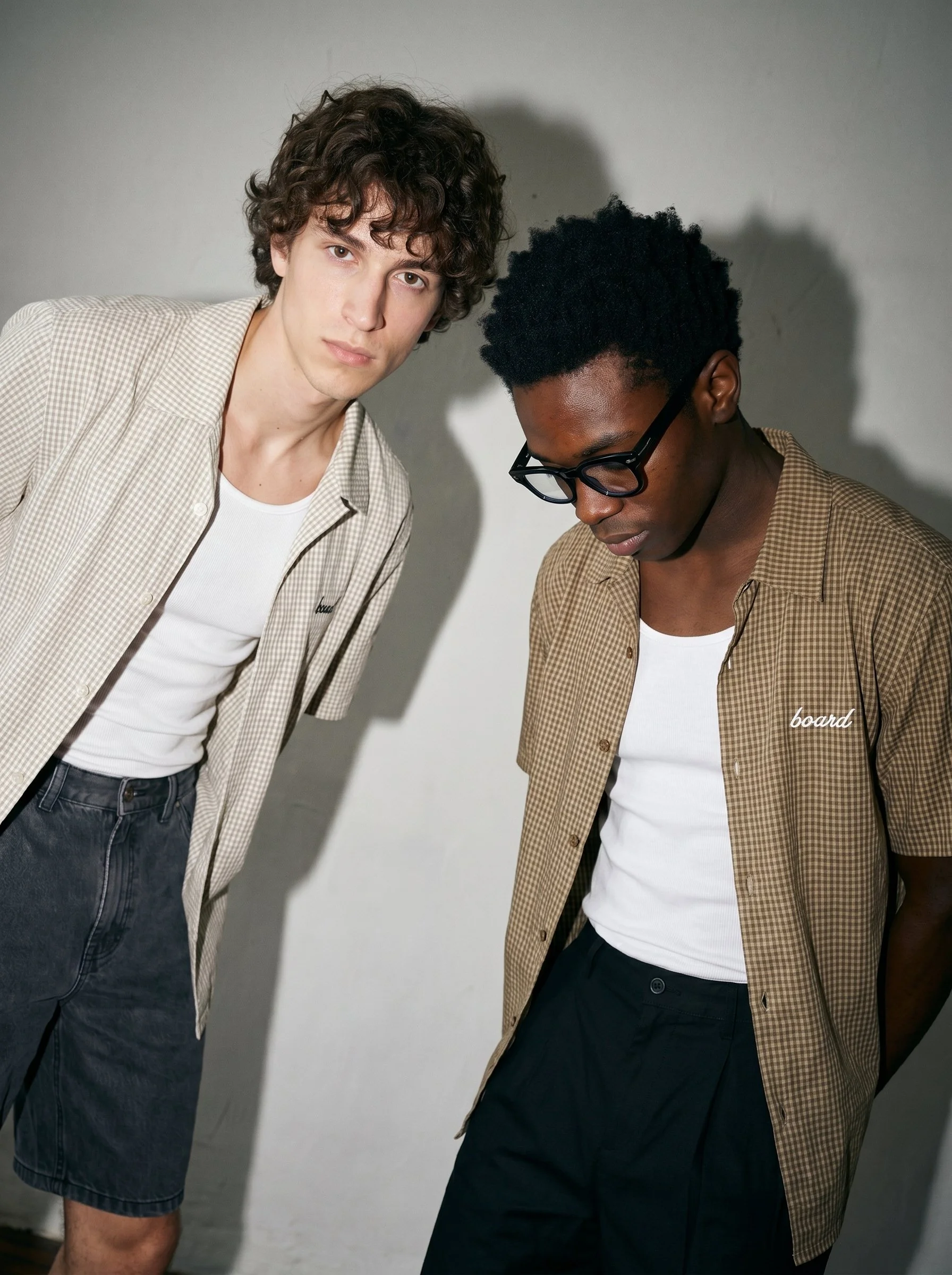Two young men stand close together against a plain wall, both wearing beige checks shirts and white t-shirts. One with curly dark hair and glasses looking down, the other with wavy light hair staring directly at the camera.