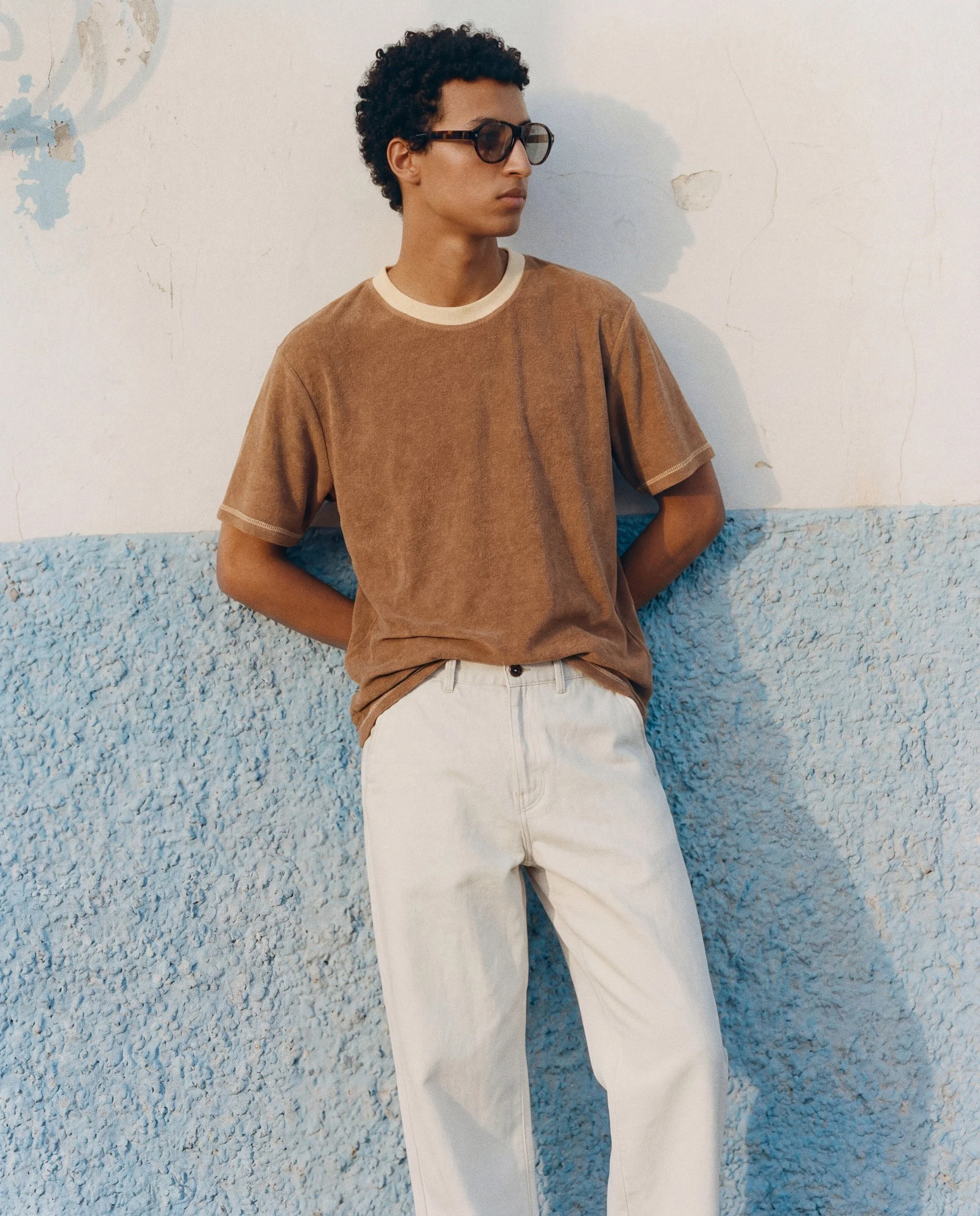 A young person with short curly hair, wearing large sunglasses, a brown t-shirt, and cream-colored pants, leaning against a textured blue and white wall.