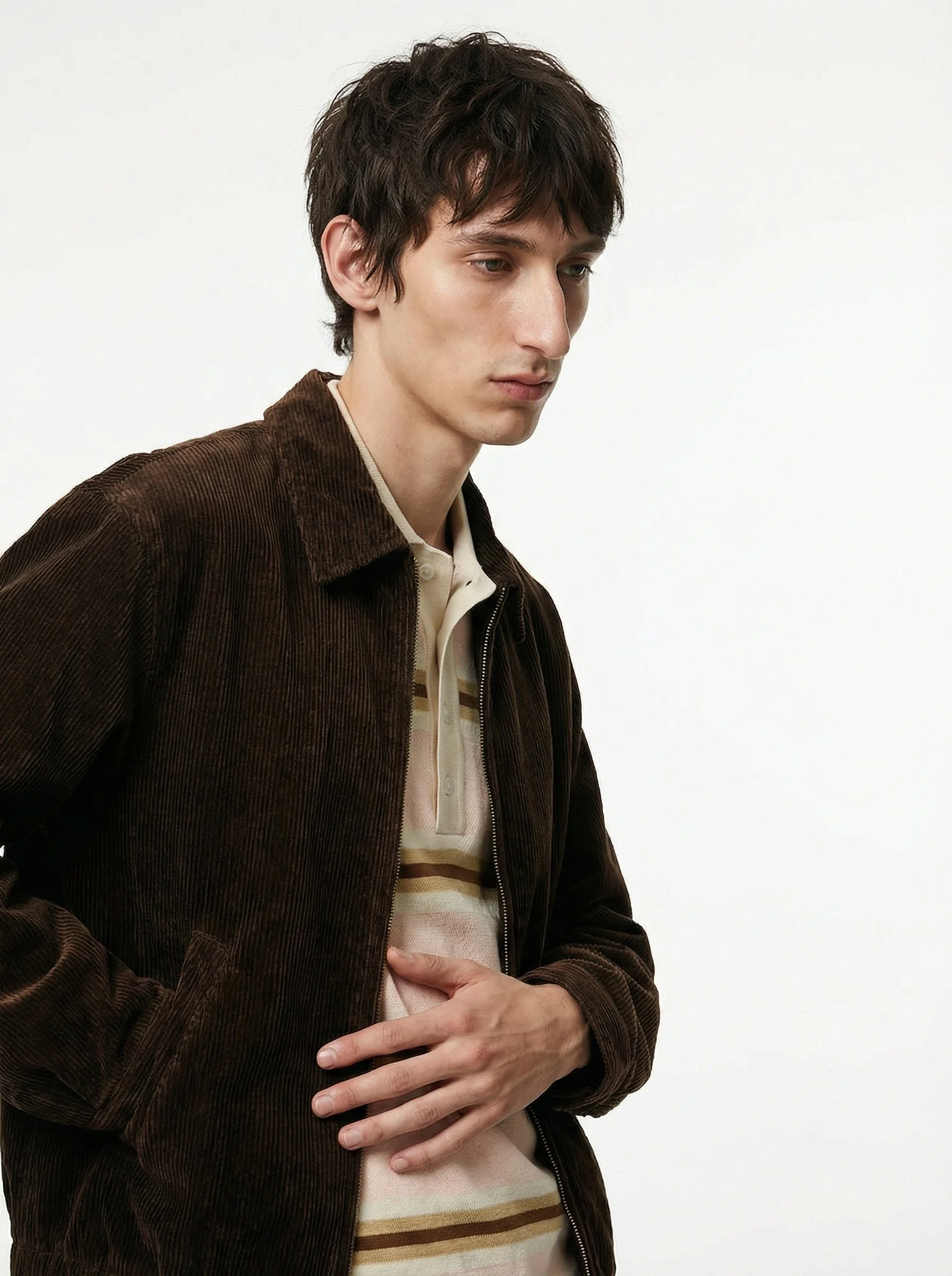 A young man with dark hair wearing a brown corduroy jacket over a beige and brown striped shirt, standing against a plain white background, with his head slightly tilted and his eyes looking downward.