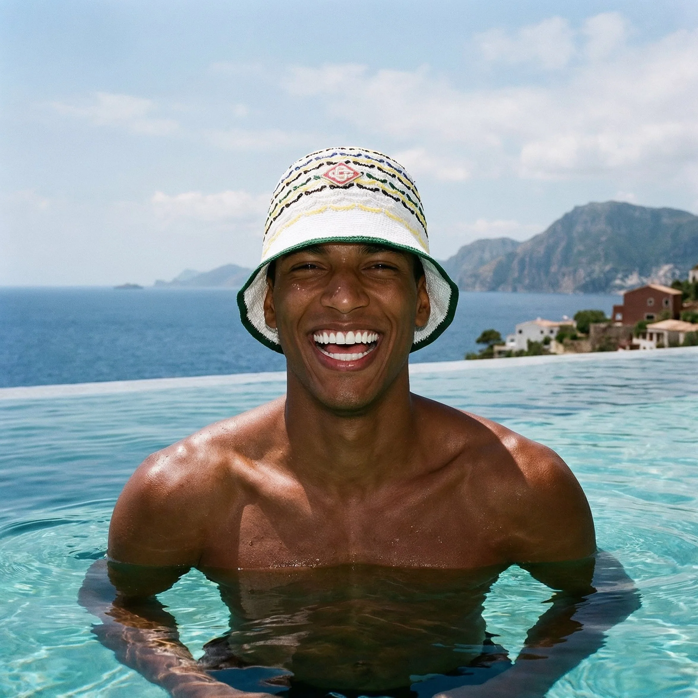 A smiling man with dark skin, wearing a colorful woven hat, in a swimming pool with scenic mountains and houses in the background. AI fashion editorial image. Casablanca.