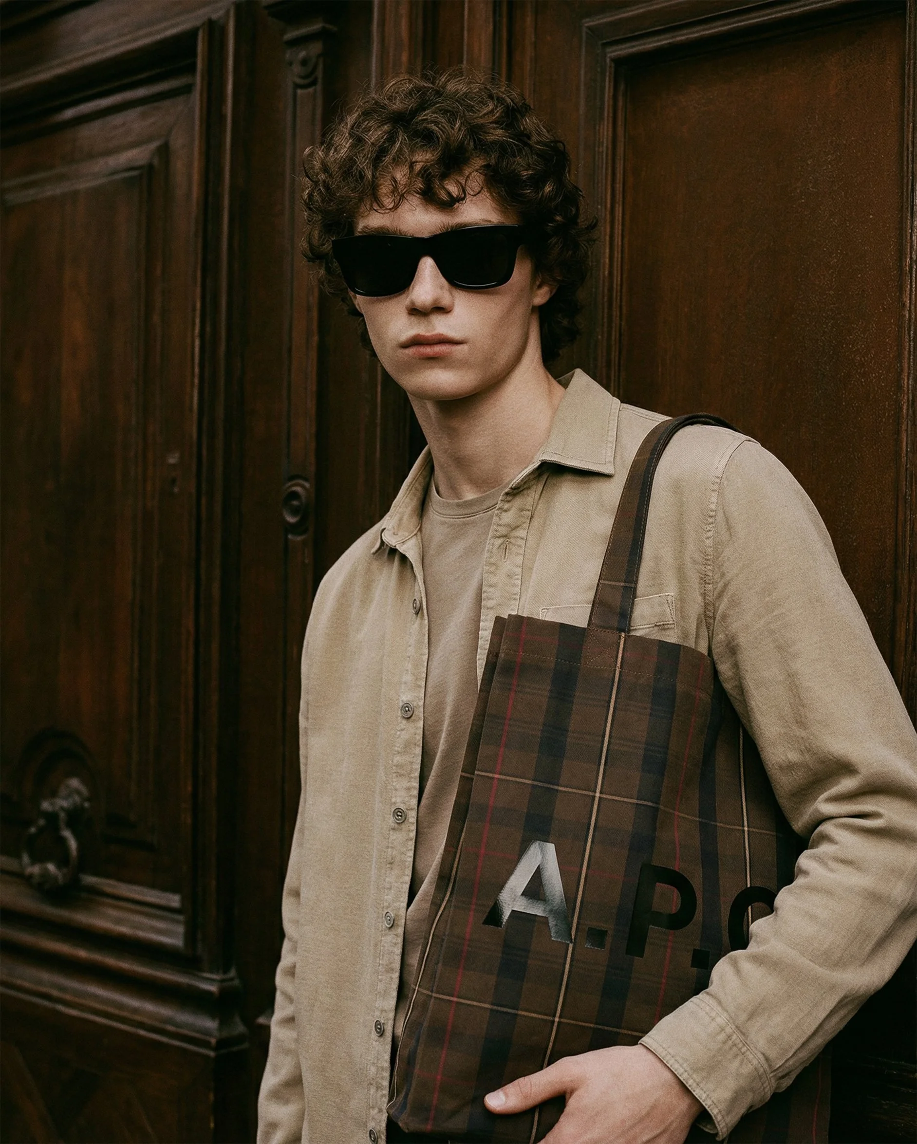 A young man with curly hair wearing black sunglasses, a beige shirt, and carrying a plaid tote bag standing in front of wooden doors.