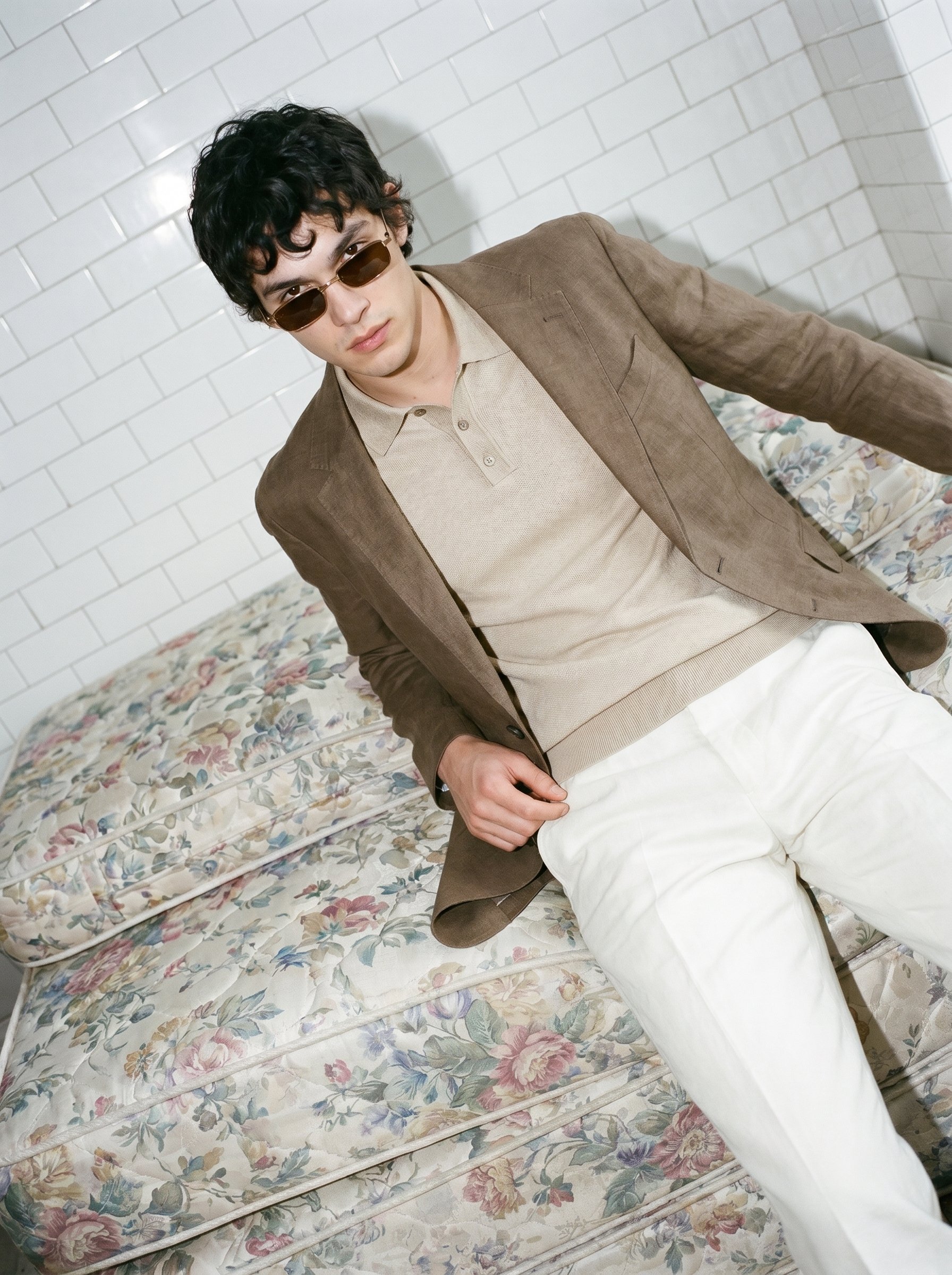 A young man with curly dark hair and sunglasses, wearing a beige polo shirt and a brown blazer, sitting on a floral-patterned mattress against a white brick wall.