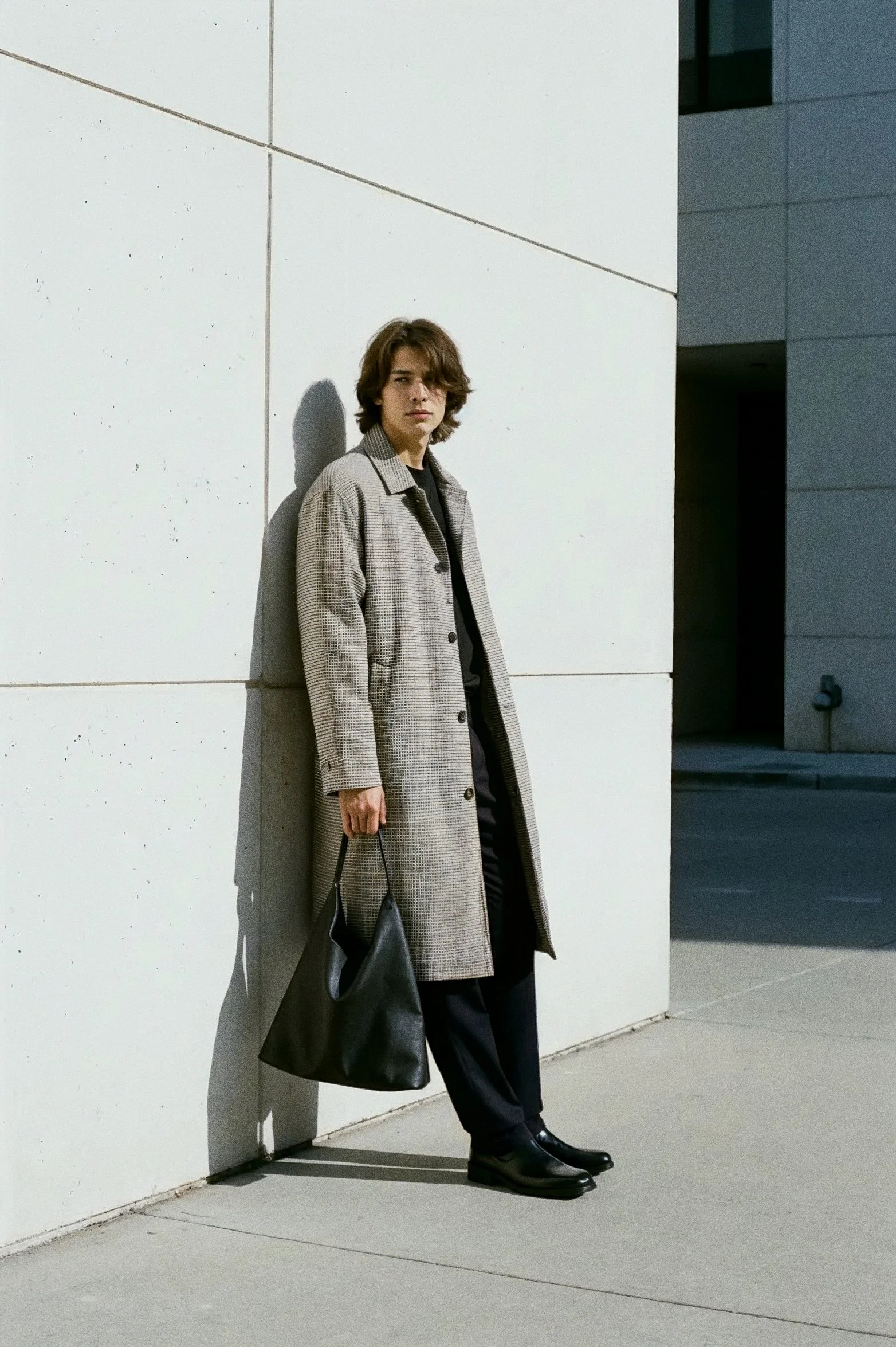 A young man with shoulder-length brown hair wearing a long checkered coat, black pants, and black boots standing against a white concrete wall, holding a black bag, with a modern building in the background.