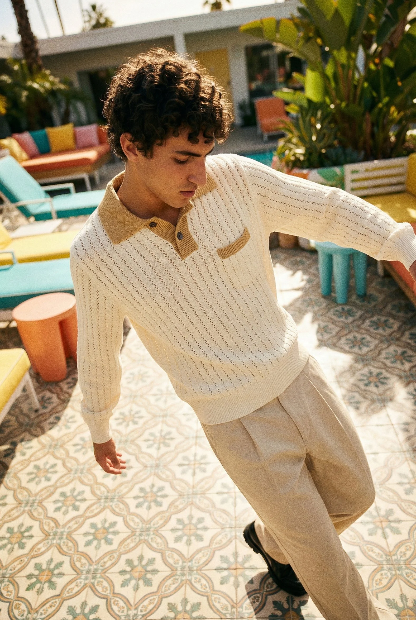 A person with curly hair wearing a cream-colored sweater and beige pants standing outdoors on a patterned tile floor, with a colorful seating area and greenery in the background.