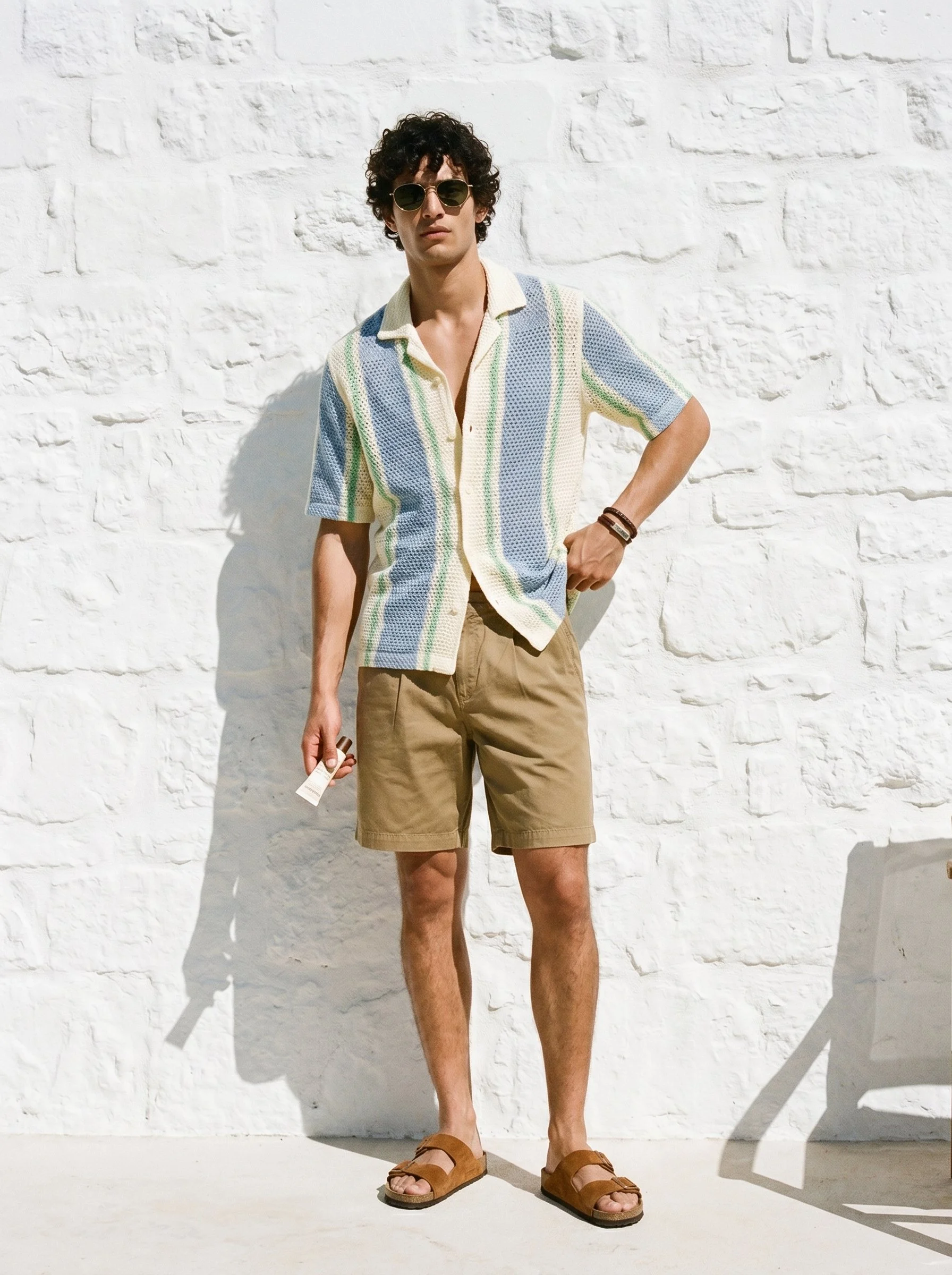Young man with curly dark hair wearing sunglasses, a short-sleeve striped shirt, khaki shorts, tan sandals, standing against a white brick wall holding a small bottle.