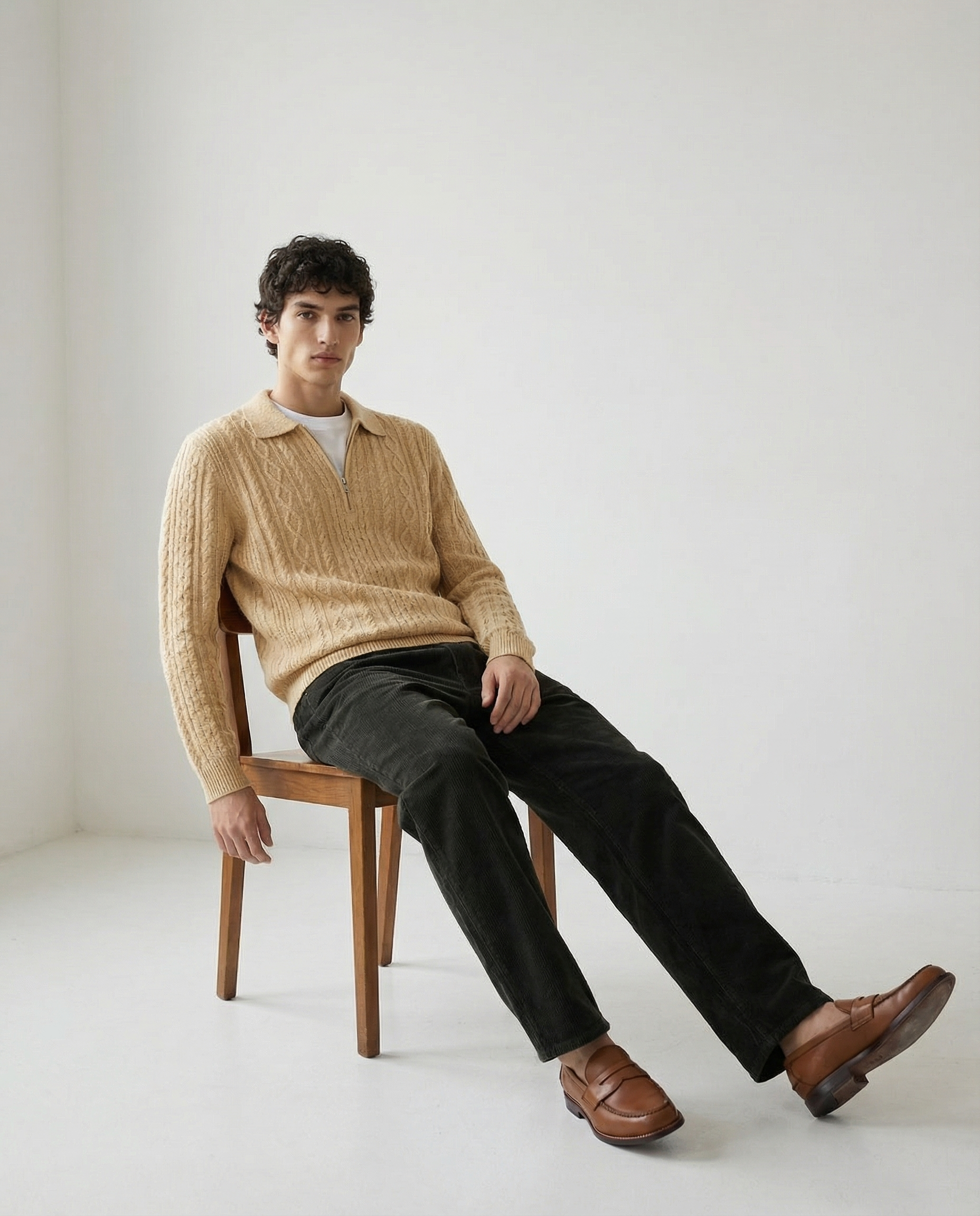 A young man with curly dark hair sitting on a wooden chair in a plain white room, wearing a beige cable knit sweater, black pants, and brown loafers.