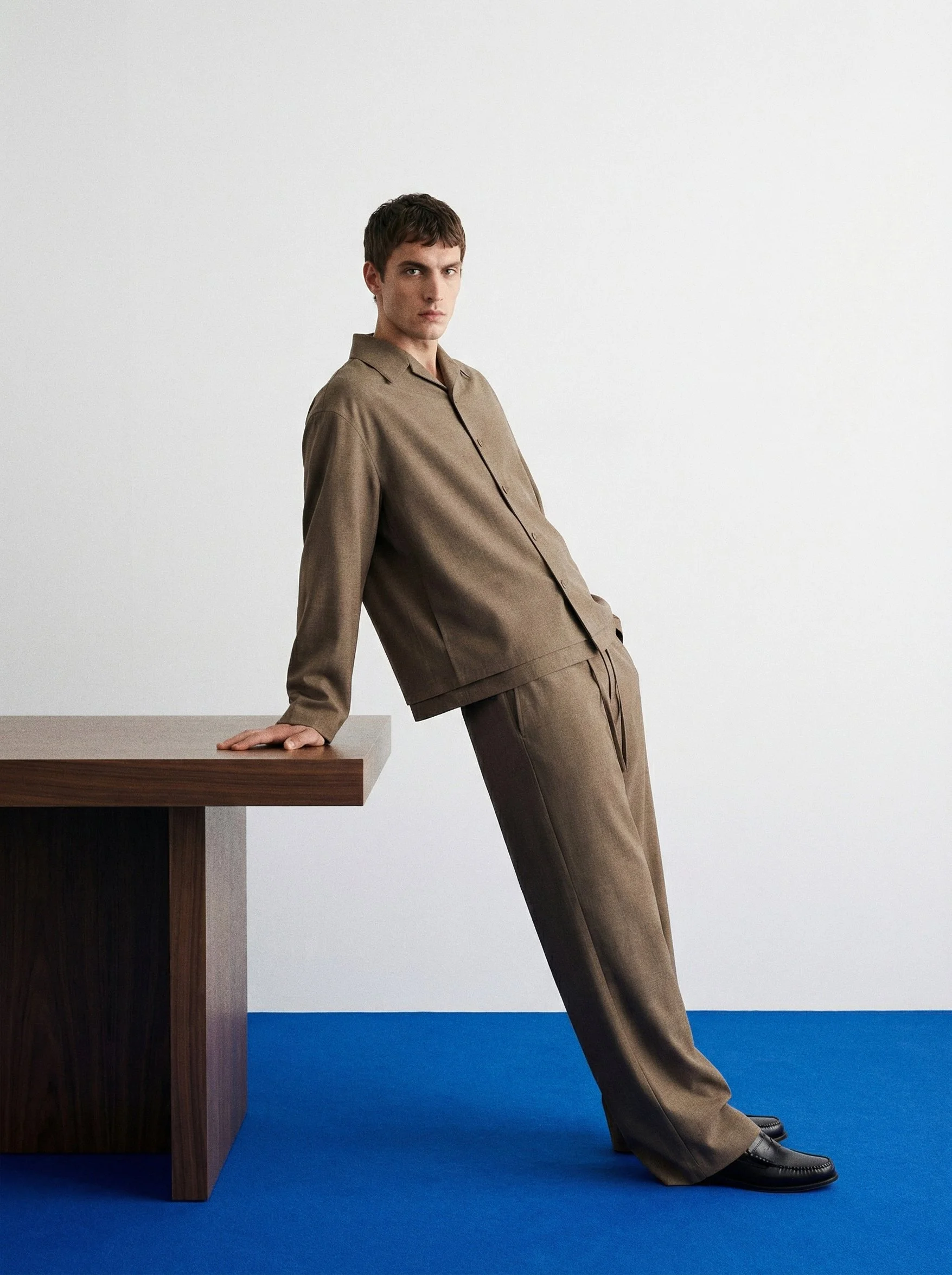 A young man with dark hair, dressed in a loose brown suit, leaning back against a plain white wall. He is resting his hand on a wooden table and wearing black shoes. AI fashion editorial.