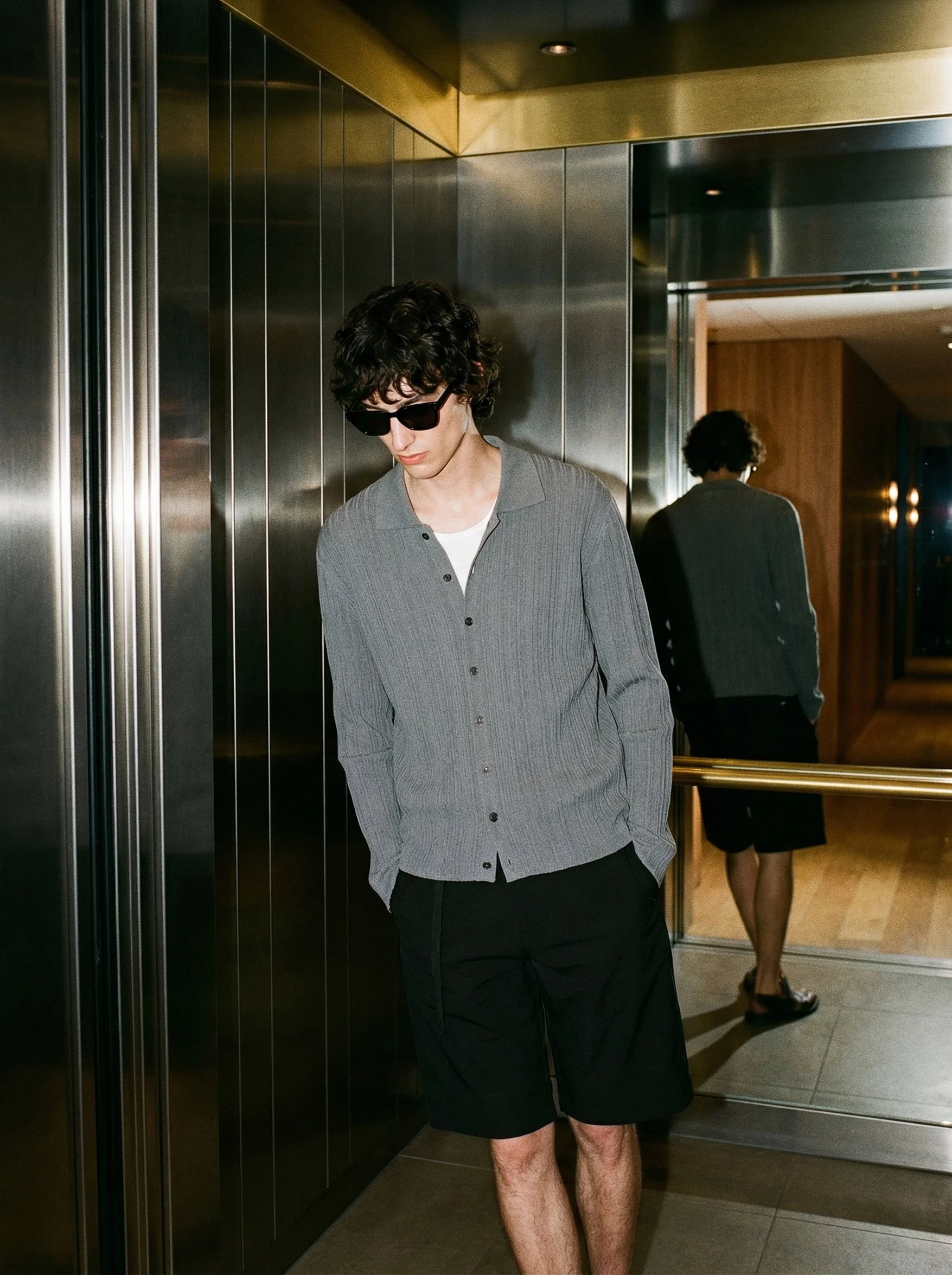 A young man with curly black hair, wearing sunglasses, a gray button-up shirt, and black shorts, standing in front of a mirrored elevator door in an indoor setting.