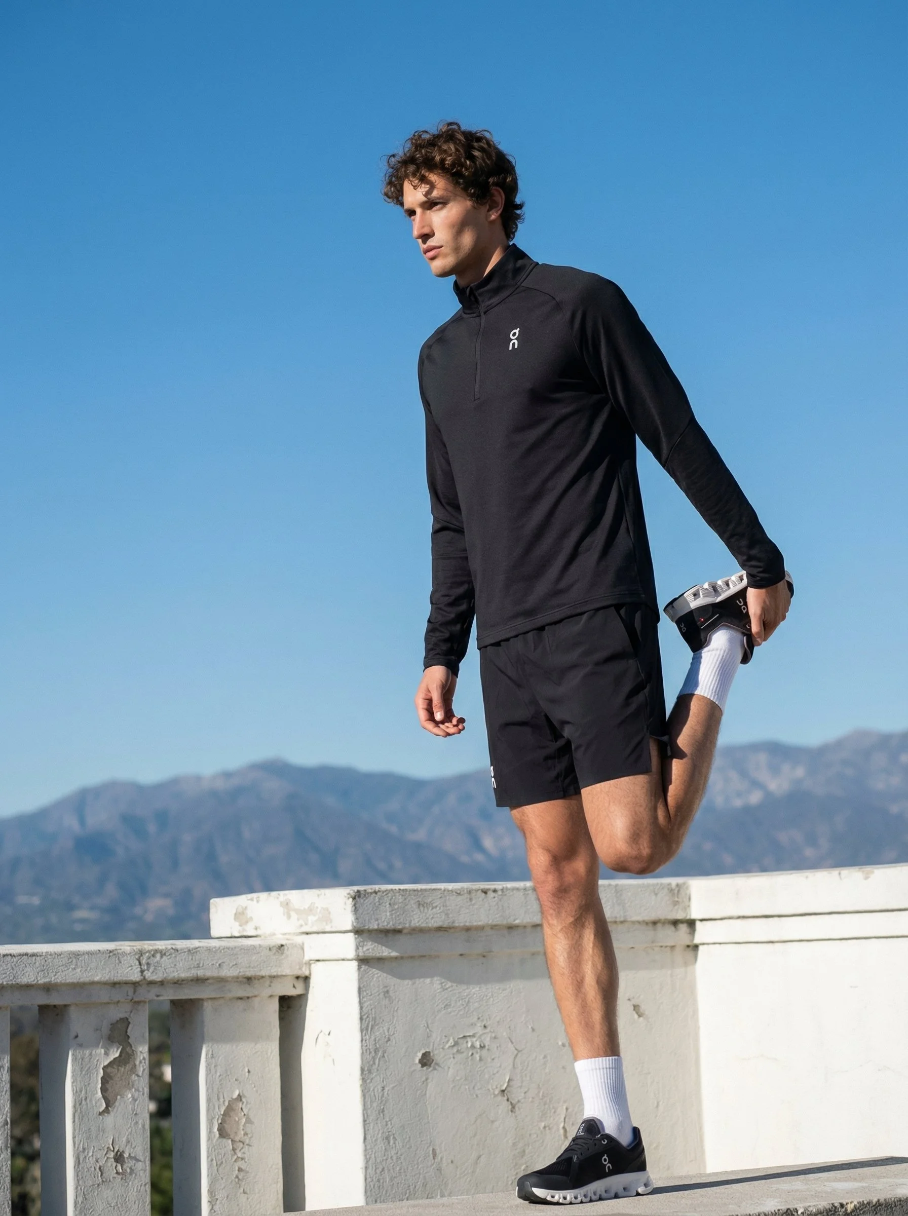 A young man stretching outdoors on a sunny day, wearing black athletic clothing, black running shoes, and white socks, with mountains in the background.