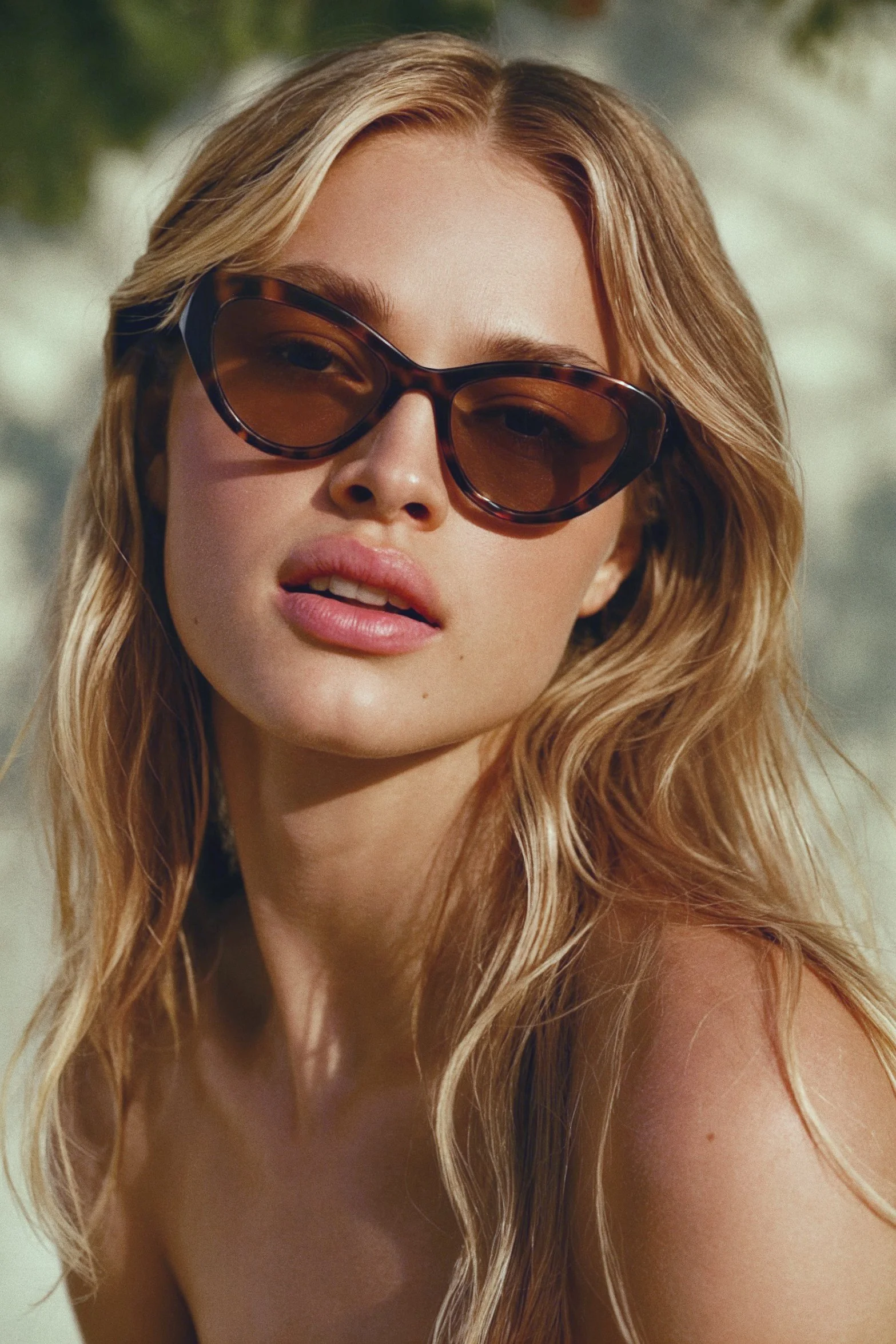 Close-up of a young woman with long, wavy blonde hair wearing large, tortoise-shell sunglasses, outdoors with blurred greenery in the background.