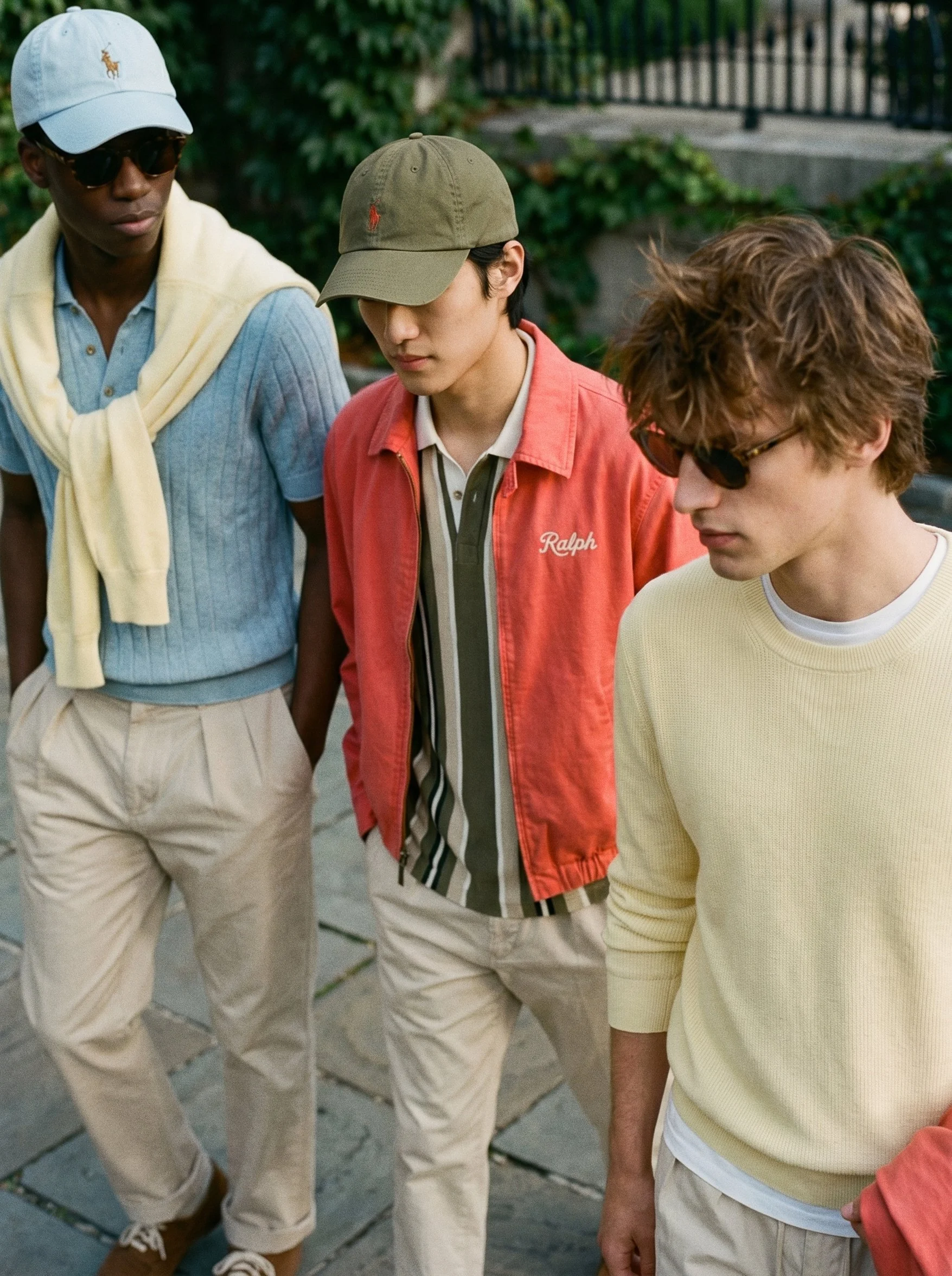 Three young men standing closely together outdoors, wearing casual and colorful clothing, with green foliage and a fence in the background.