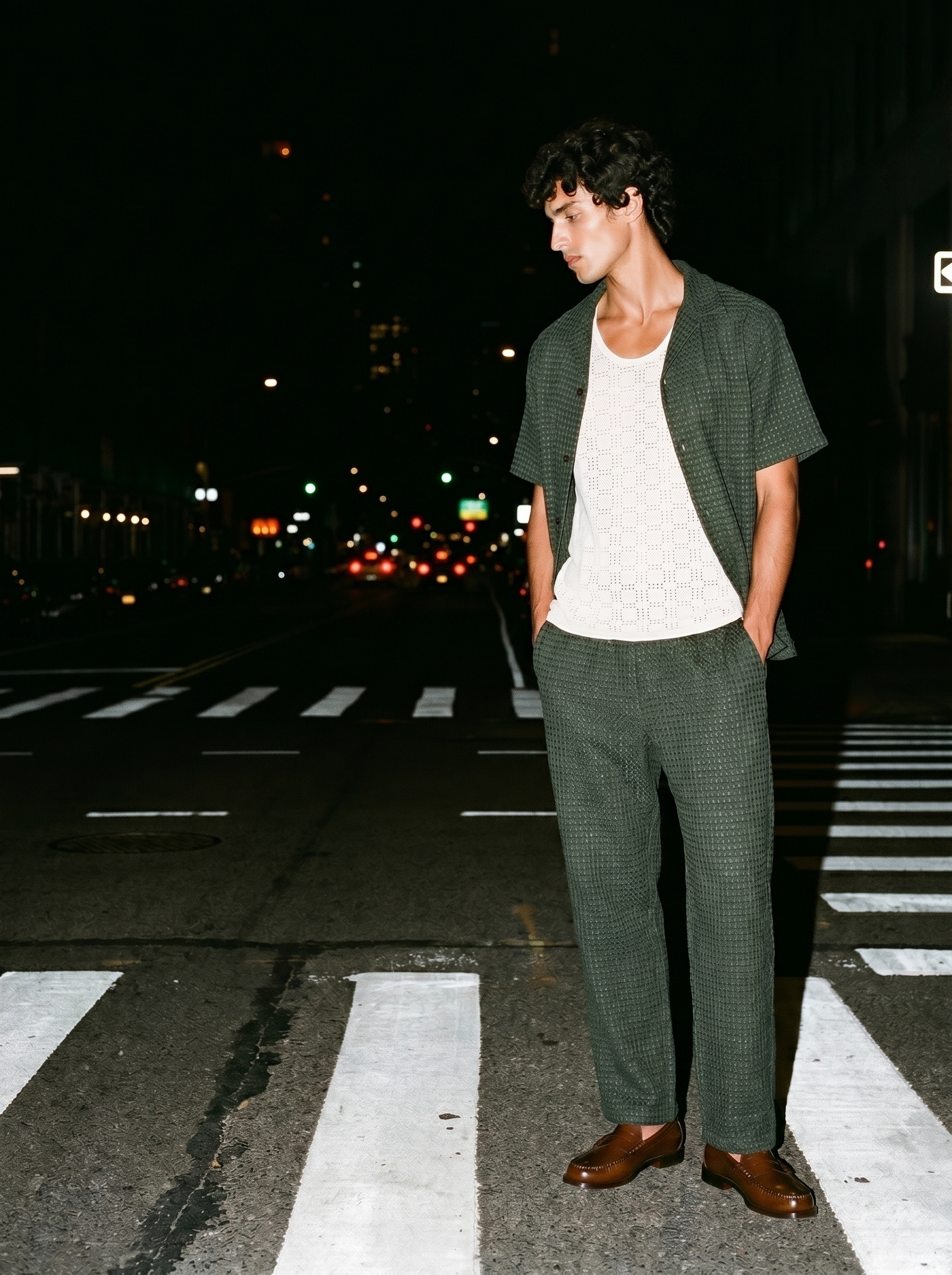 A man wearing a green checked suit and brown shoes is standing on a crosswalk at night with city lights in the background.