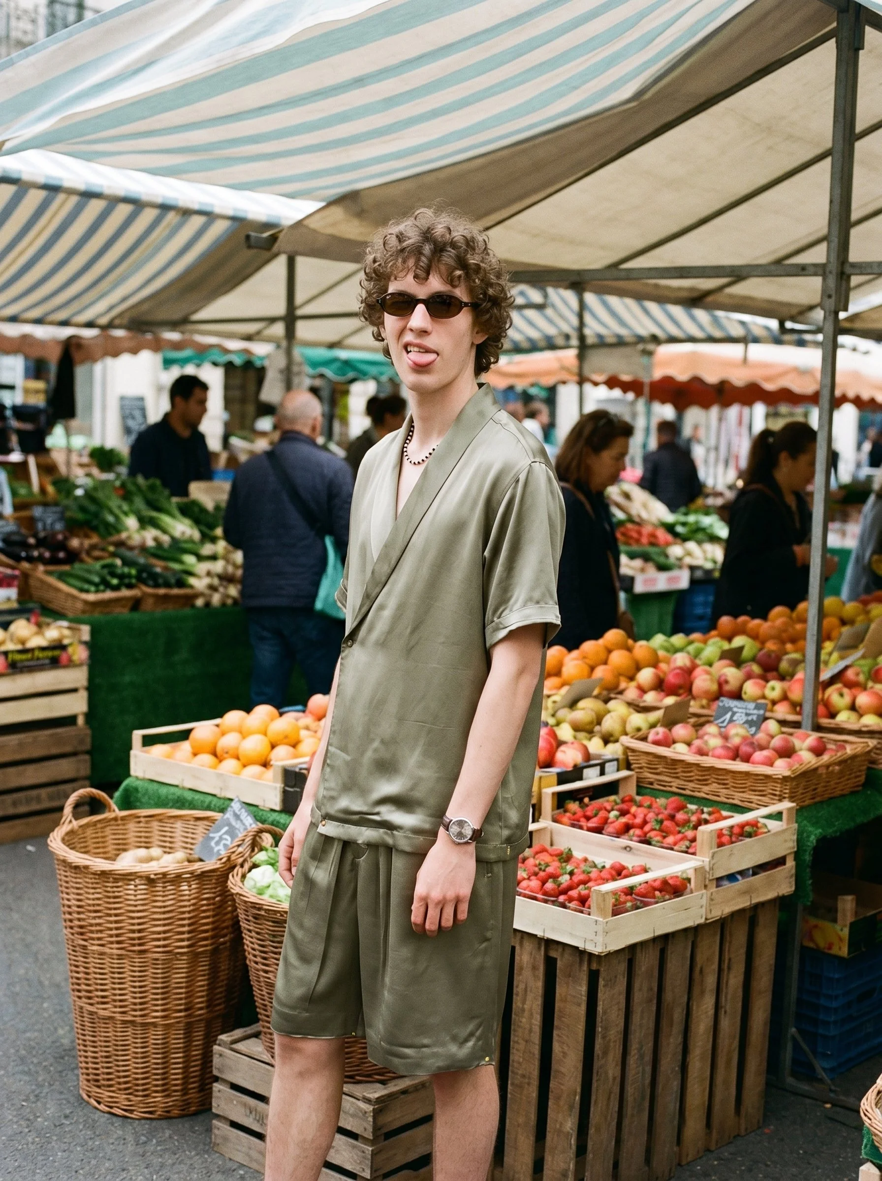 A young man in sunglasses and a beige outfit sticking out his tongue at a farmer's market. The market has fruit and vegetable stalls with striped umbrellas.