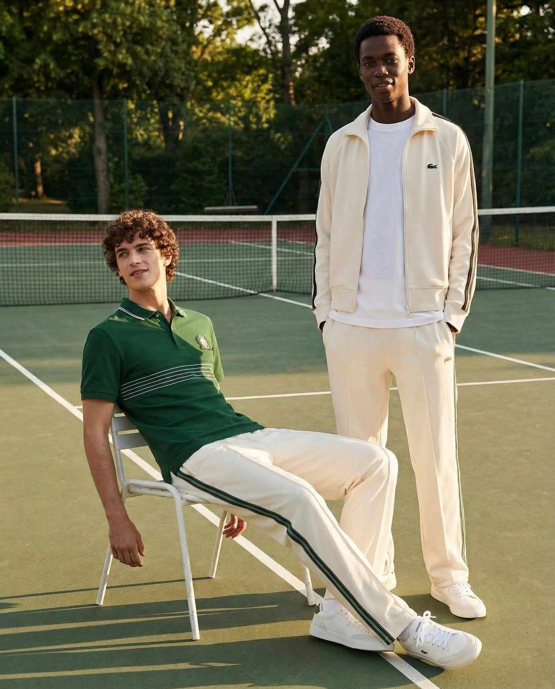 Two young men on a tennis court during sunset. One sitting on a chair, wearing a green Lacoste polo and white pants, the other standing, wearing a cream-colored athletic jacket and matching pants, and white sneakers.