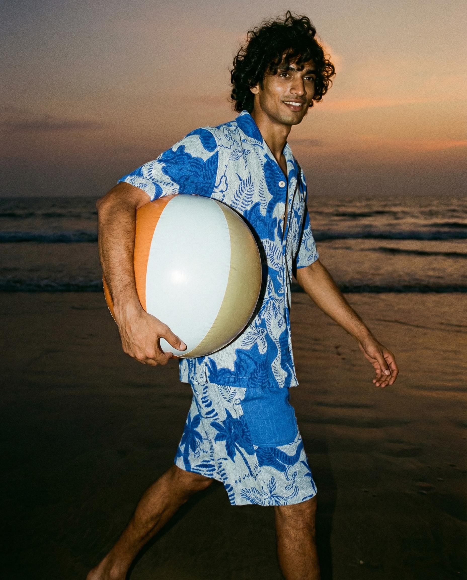 A man with curly dark hair wearing a blue Hawaiian shirt and blue shorts is walking on a beach at sunset, holding a surfboard.  AI fashion editorial image.Polo Ralph Laurent.