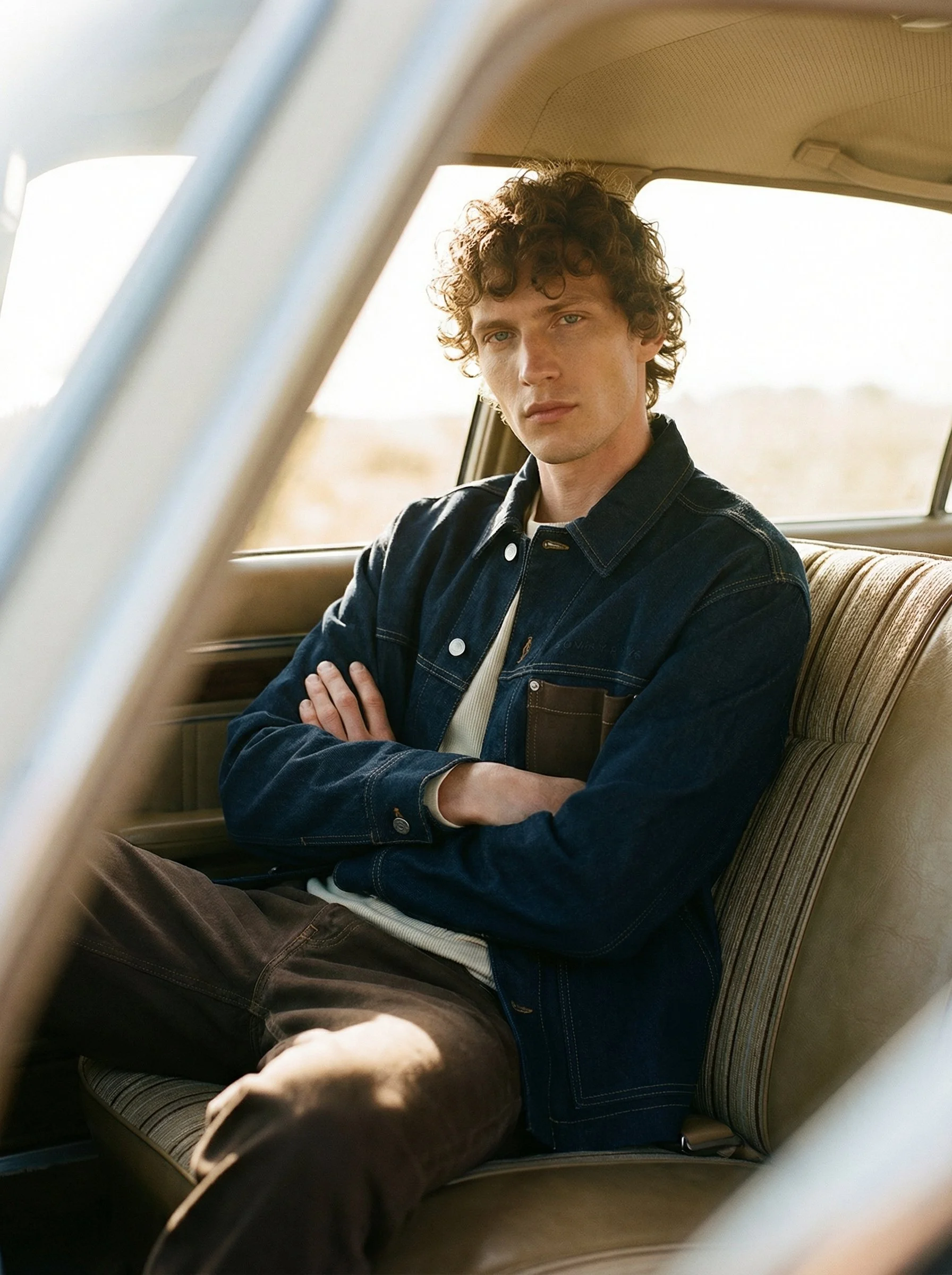 A young man with curly hair sitting in a car with his arms crossed, wearing a dark denim jacket and brown pants, looking directly at the camera.