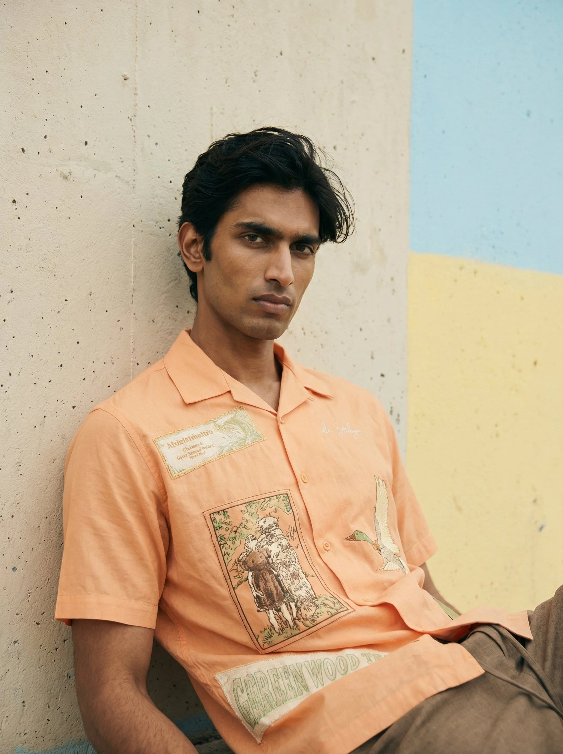 A young man with dark hair, wearing a peach-colored shirt with patches, sitting against a beige wall with colorful geometric shapes, looking at the camera.