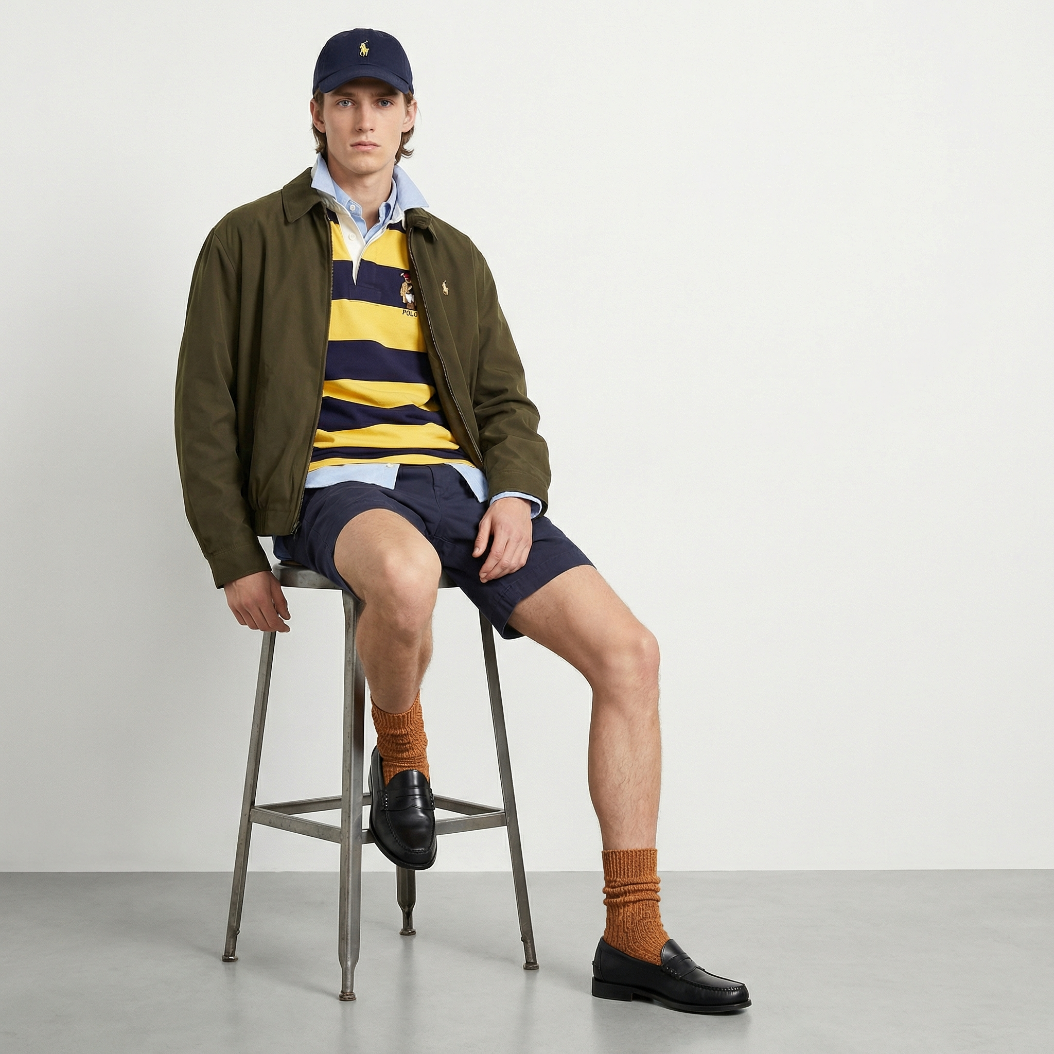 A young male model sitting on a metal stool against a plain light gray background, wearing a navy blue baseball cap with a yellow logo, a yellow and navy striped polo shirt, a green jacket, navy shorts, orange socks, and black shoes.