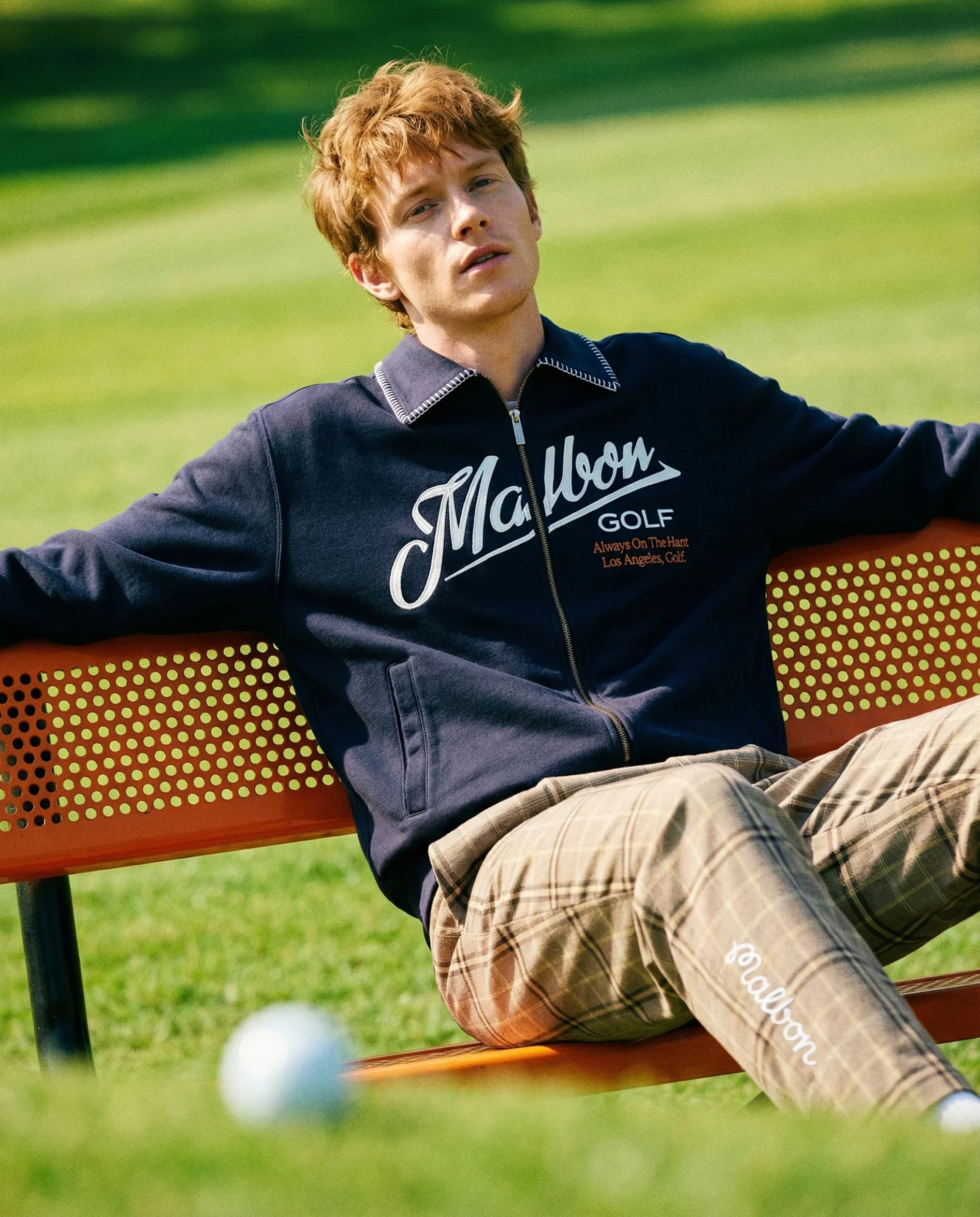 A young man with red hair wearing a navy blue jacket and beige plaid shorts sits on a red park bench in a grassy outdoor area, looking at the camera with a relaxed expression.