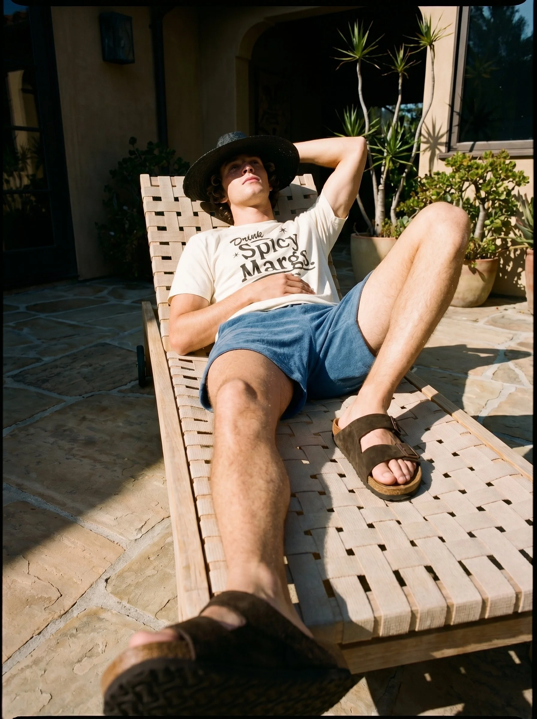 A young man is lounging on a wooden outdoor chair on a patio, wearing a large black hat, a beige T-shirt with text, blue shorts, and sandals. He is resting with one arm behind his head and the other hand on his stomach. The patio has potted plants an