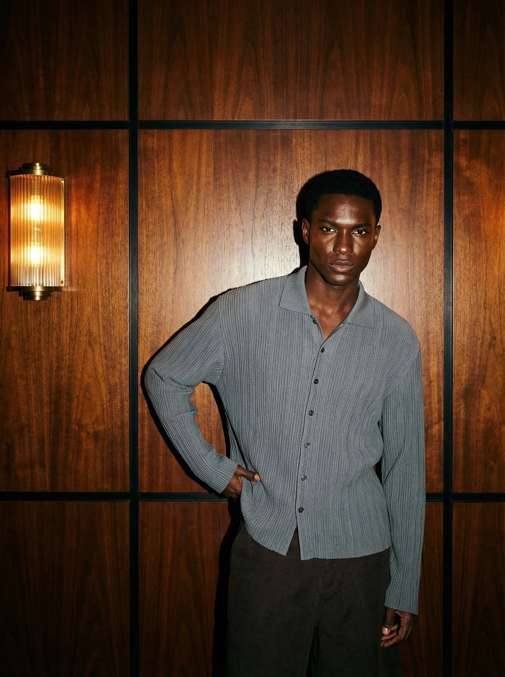 A young man with dark skin and short black hair stands against a wooden paneled wall. He is wearing a gray striped button-up shirt and dark pants. His left hand is placed on his hip, and he is looking directly at the camera with a serious expression.
