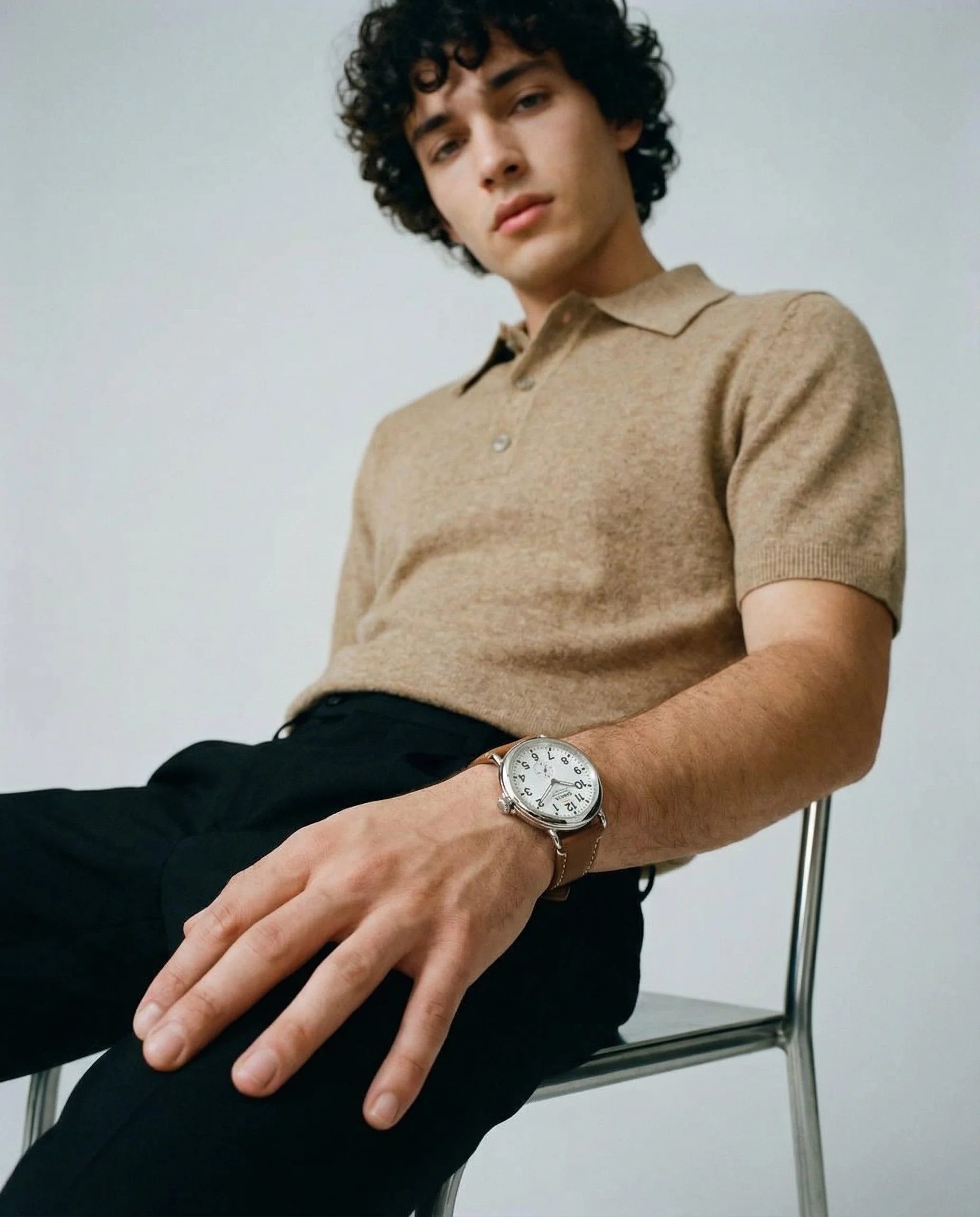 A young man with curly dark hair sitting on a metal chair against a plain white wall, wearing a beige polo shirt and a silver wristwatch, with his arm resting on the chair's armrest and his hand extended outward.