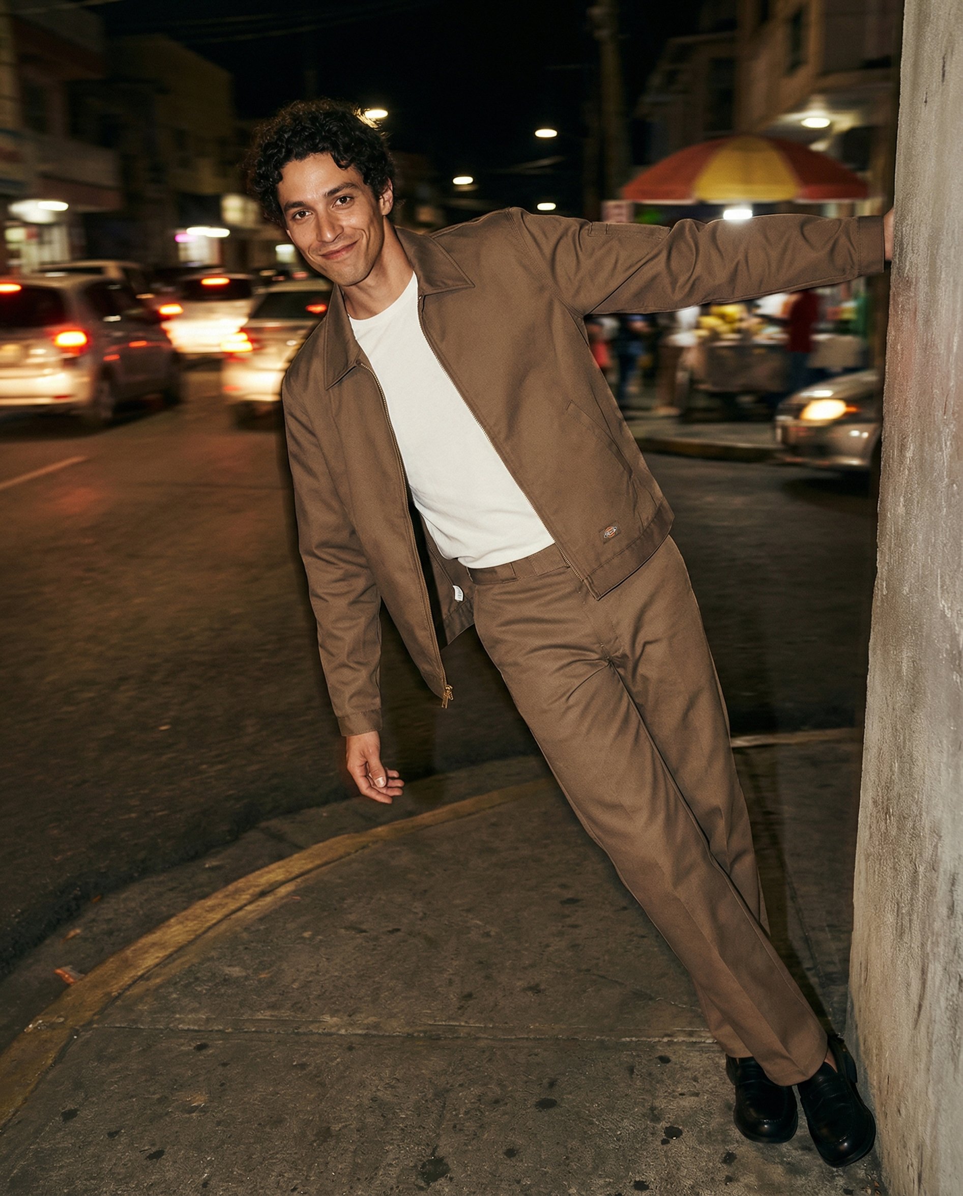 A man with curly black hair smiling and leaning against a wall on a city street at night, wearing a brown jacket, white t-shirt, brown pants, and black shoes. Cars are visible in motion background and colorful umbrellas are seen.