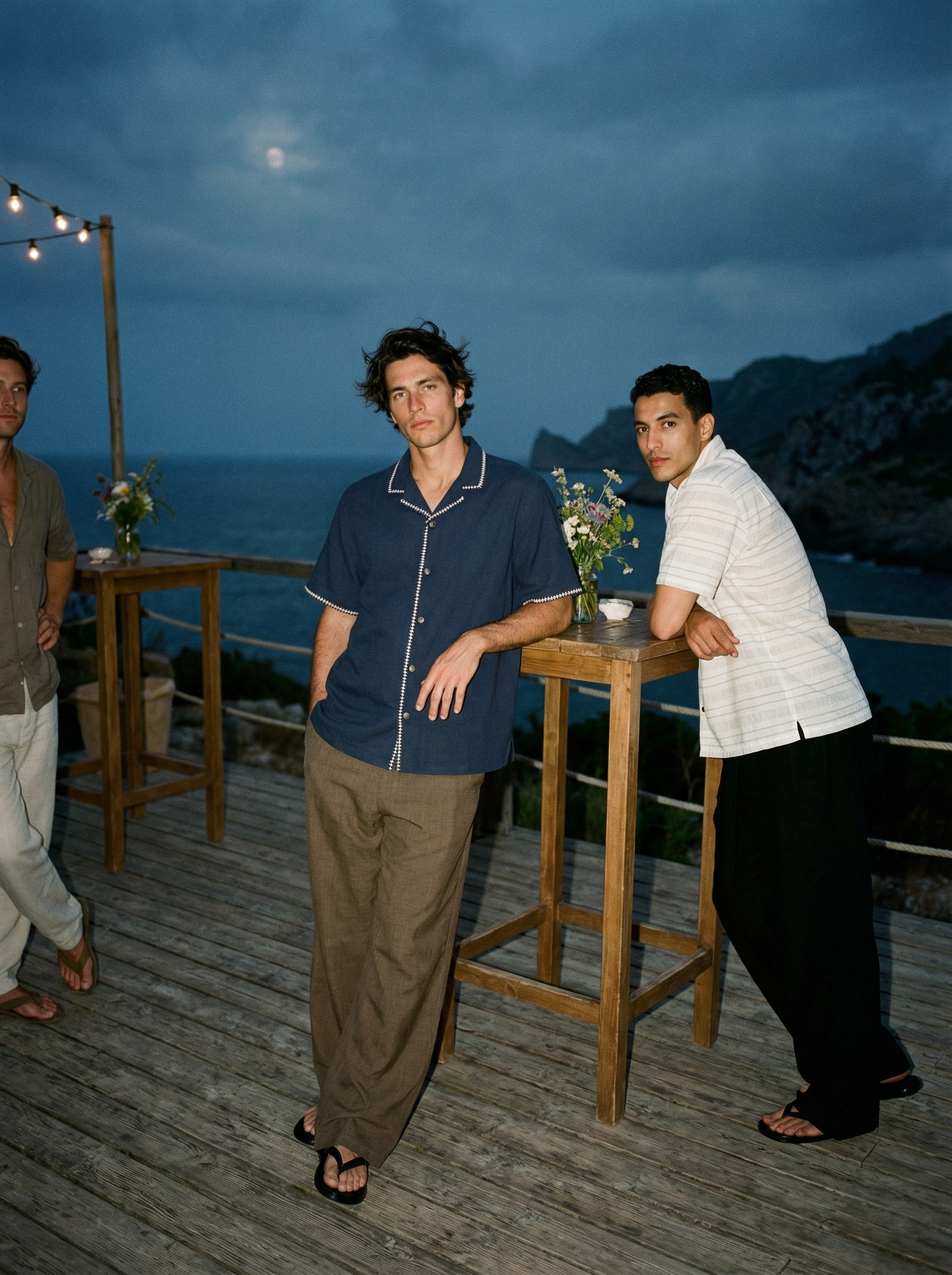 Two men standing on a wooden deck at sunset, with a view of mountains and the ocean in the background. One man is wearing a navy blue shirt, and the other is in a light-colored striped shirt. There are flower vases and string lights on the deck.