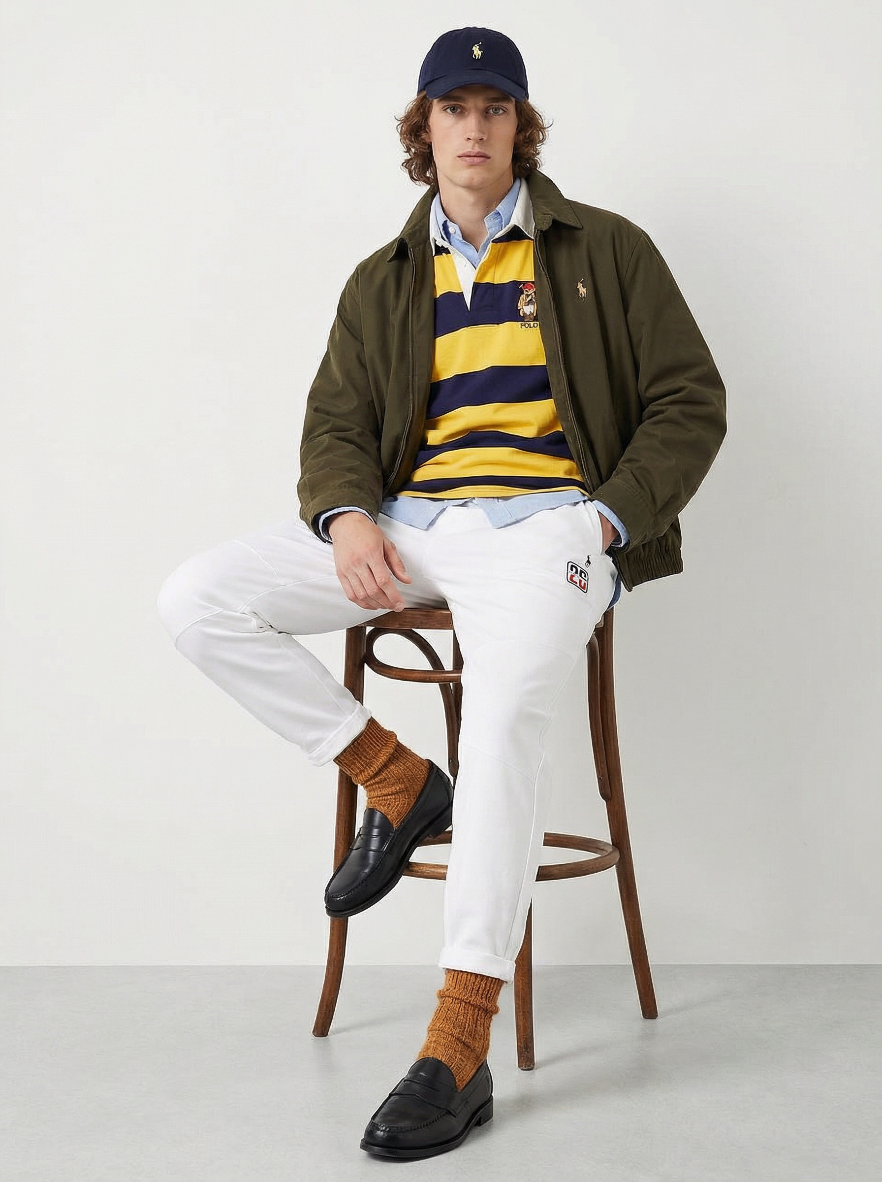 Young man sitting on a wooden chair against a white background, wearing a navy blue cap with a yellow logo, an olive green jacket, a yellow and navy striped polo shirt, white pants, brown knit socks, and black loafers.  AI fashion editorial image.