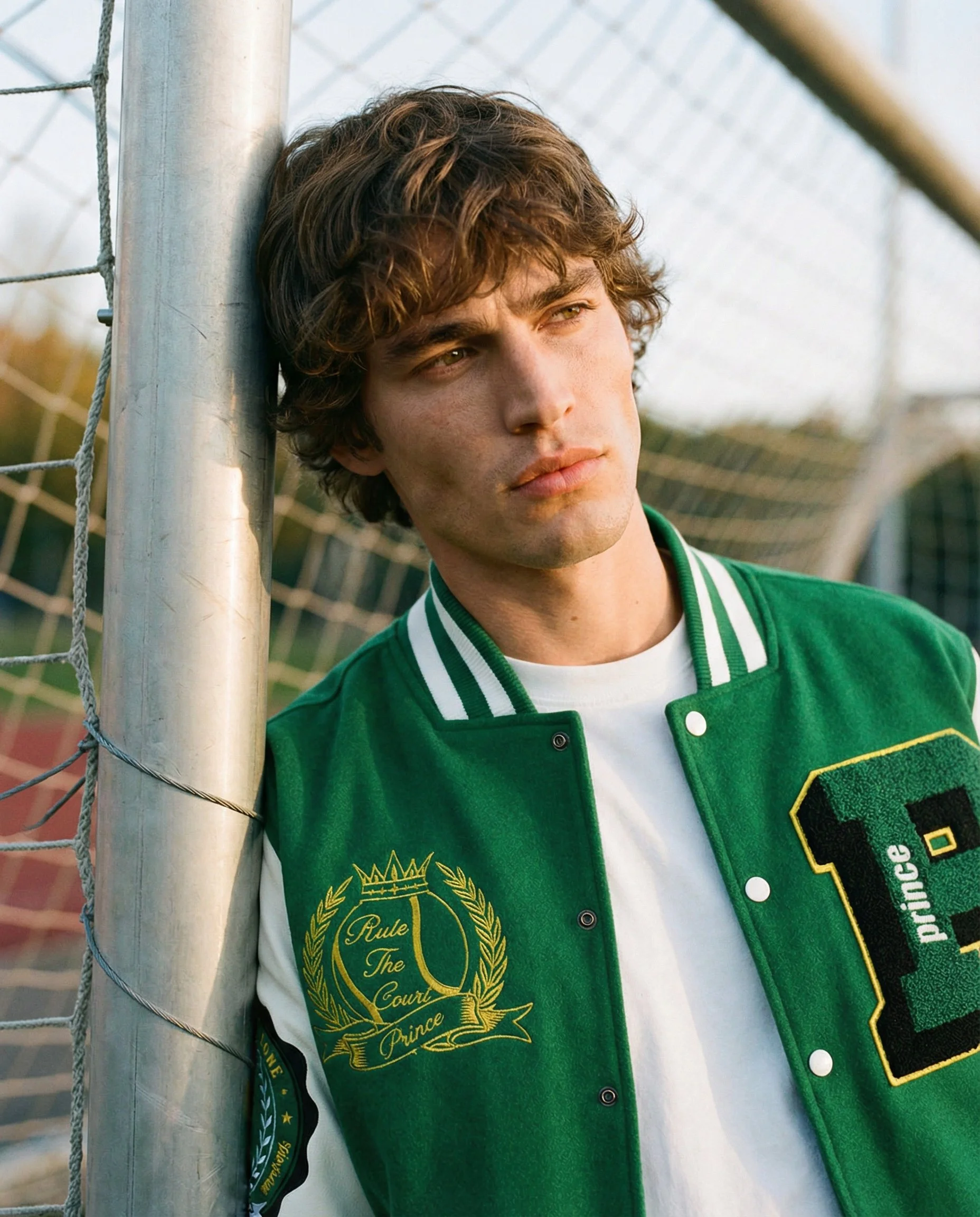 Young man with curly brown hair wearing a green varsity jacket standing next to a soccer goal post, looking off into the distance.